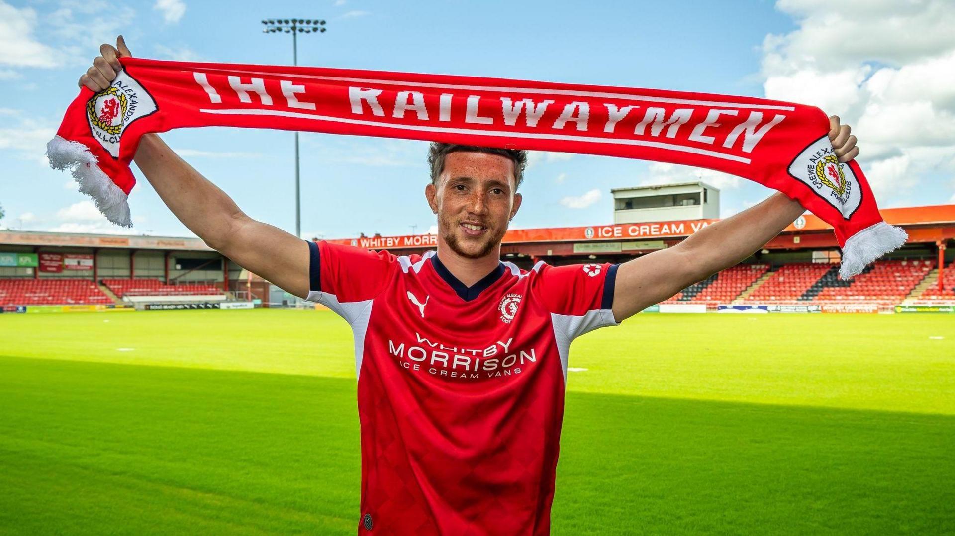 Jordan Gibson is wearing a red and white Crewe Alexandra home shirt and is holding a club scarf above his head with both arms stretched out. The scarf has Crewe's nickname The Railwaymen written on it in white letters. He is standing on the pitch at the Mornflake Stadium in bright sunshine with two stands in the background.