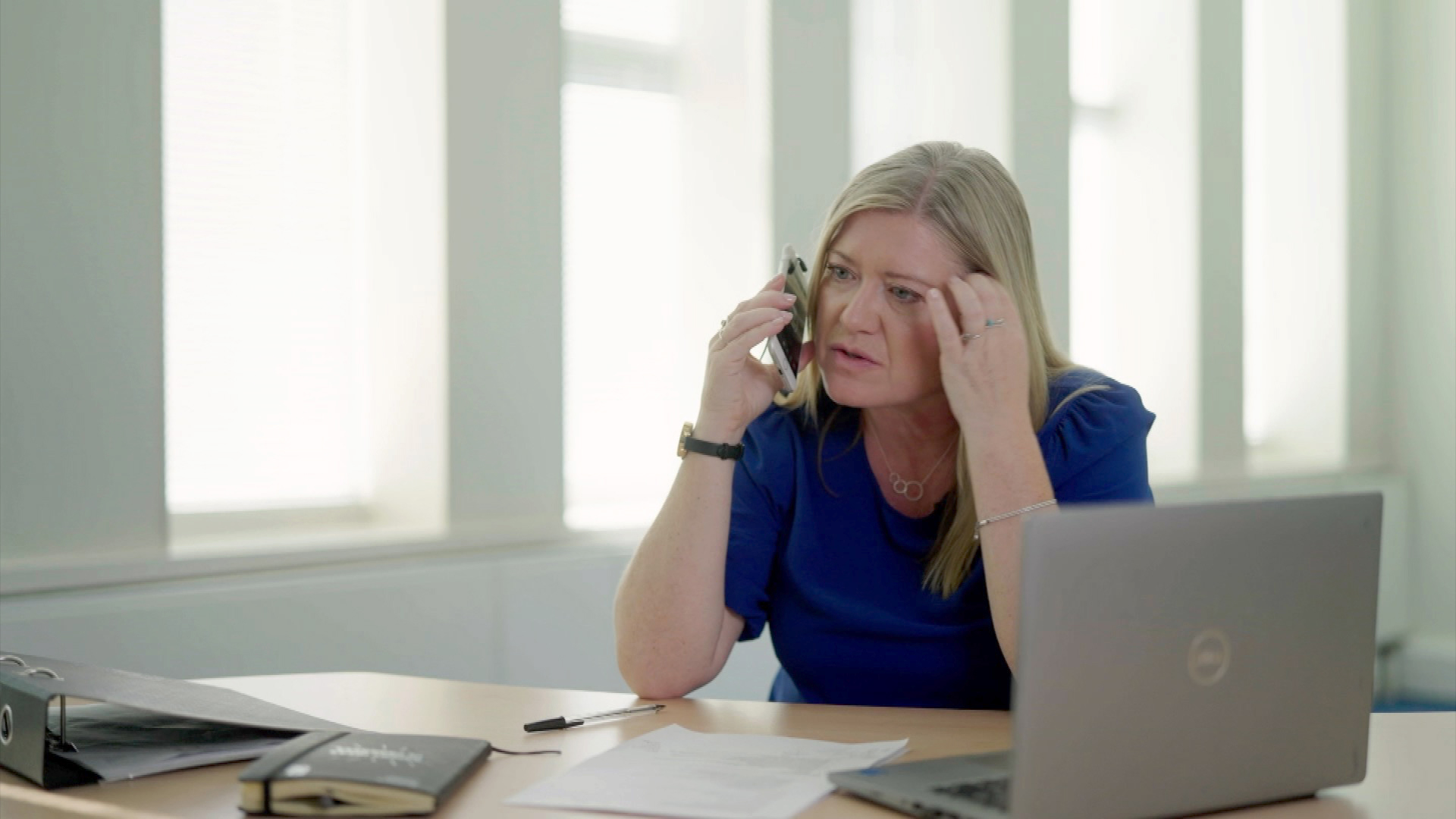 Kath Stanczyszyn in a blue top on a phone sitting in an office. There is a desk in front of her, with a laptop to her left and windows behind her