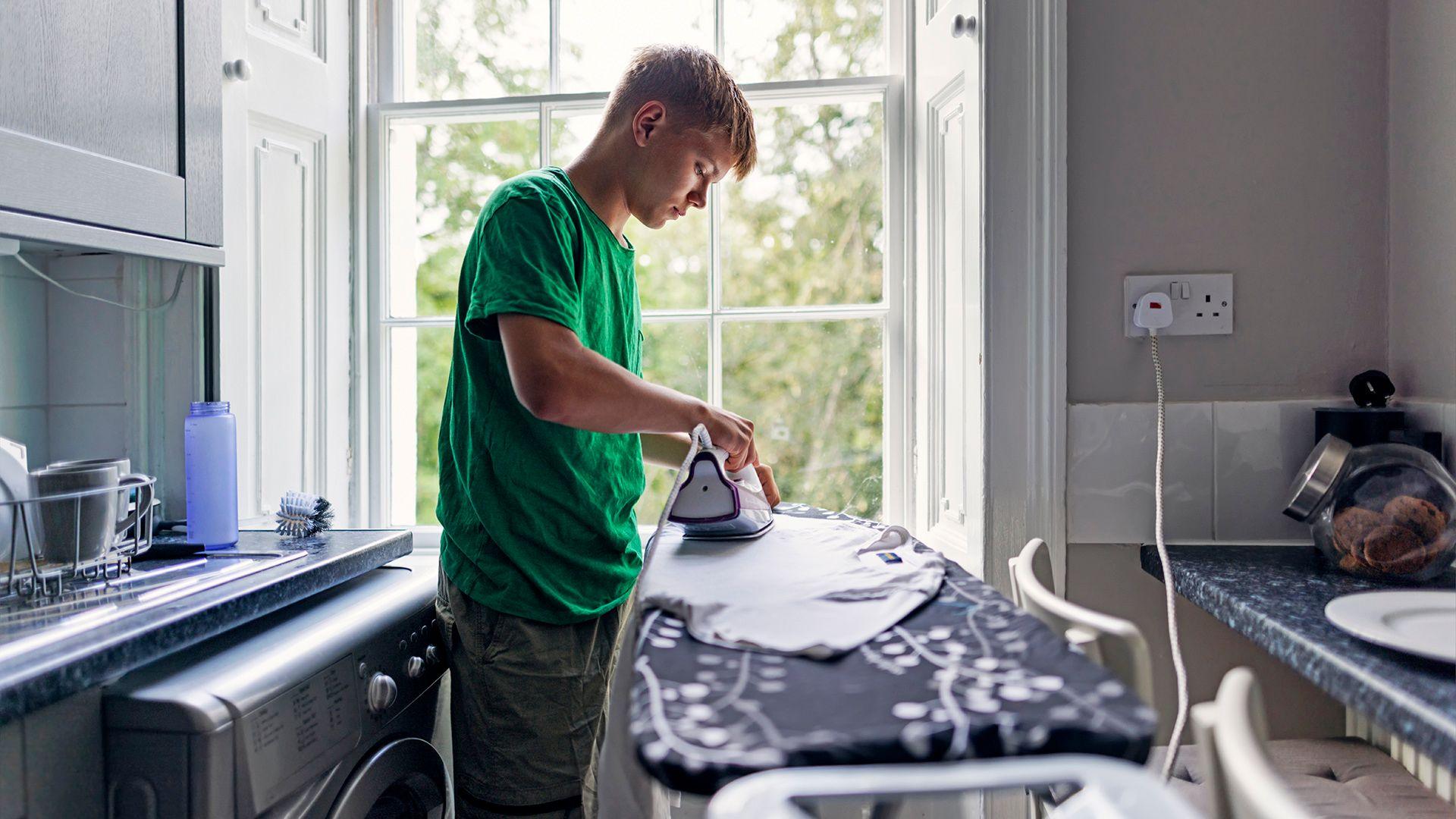 Stock photo shows a person ironing a T shirt in front of a window in their kitchen, next to a plug socket and with the washing machine behind them.