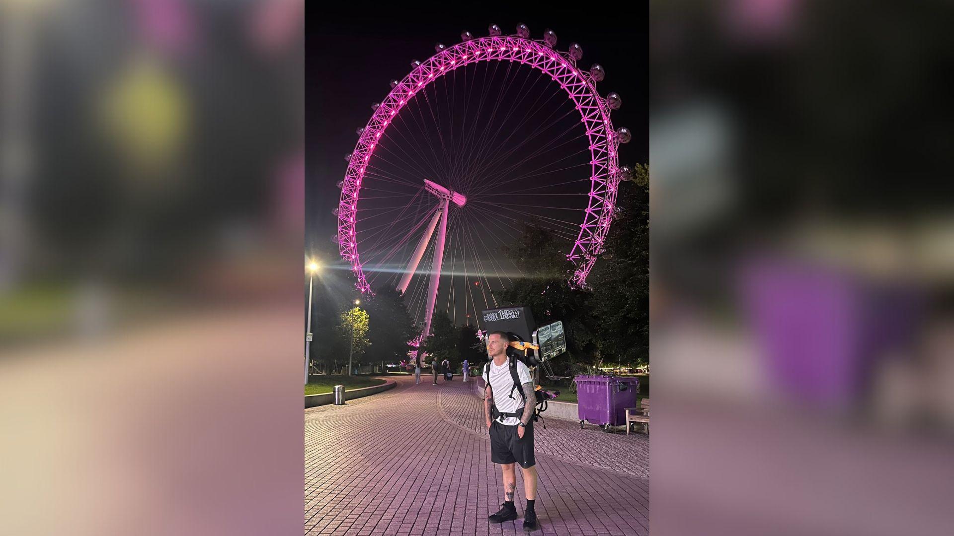 Jonathan Bradley wearing a white T-shirt and black shorts with a barber chair strapped to his back while standing in front of the London Eye. It is a large circular structure with pink lights.