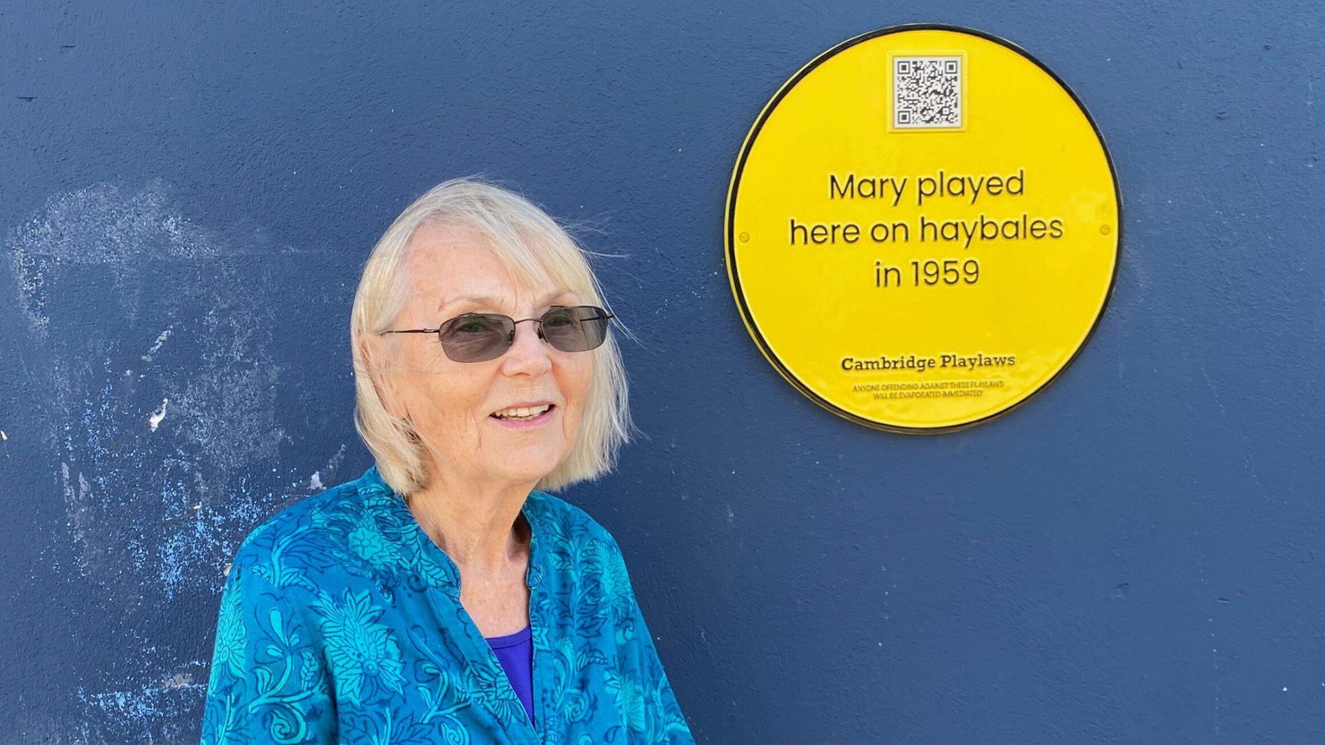 Mary standing next to her yellow plaque which says 'Mary played here on haybales in 1959".