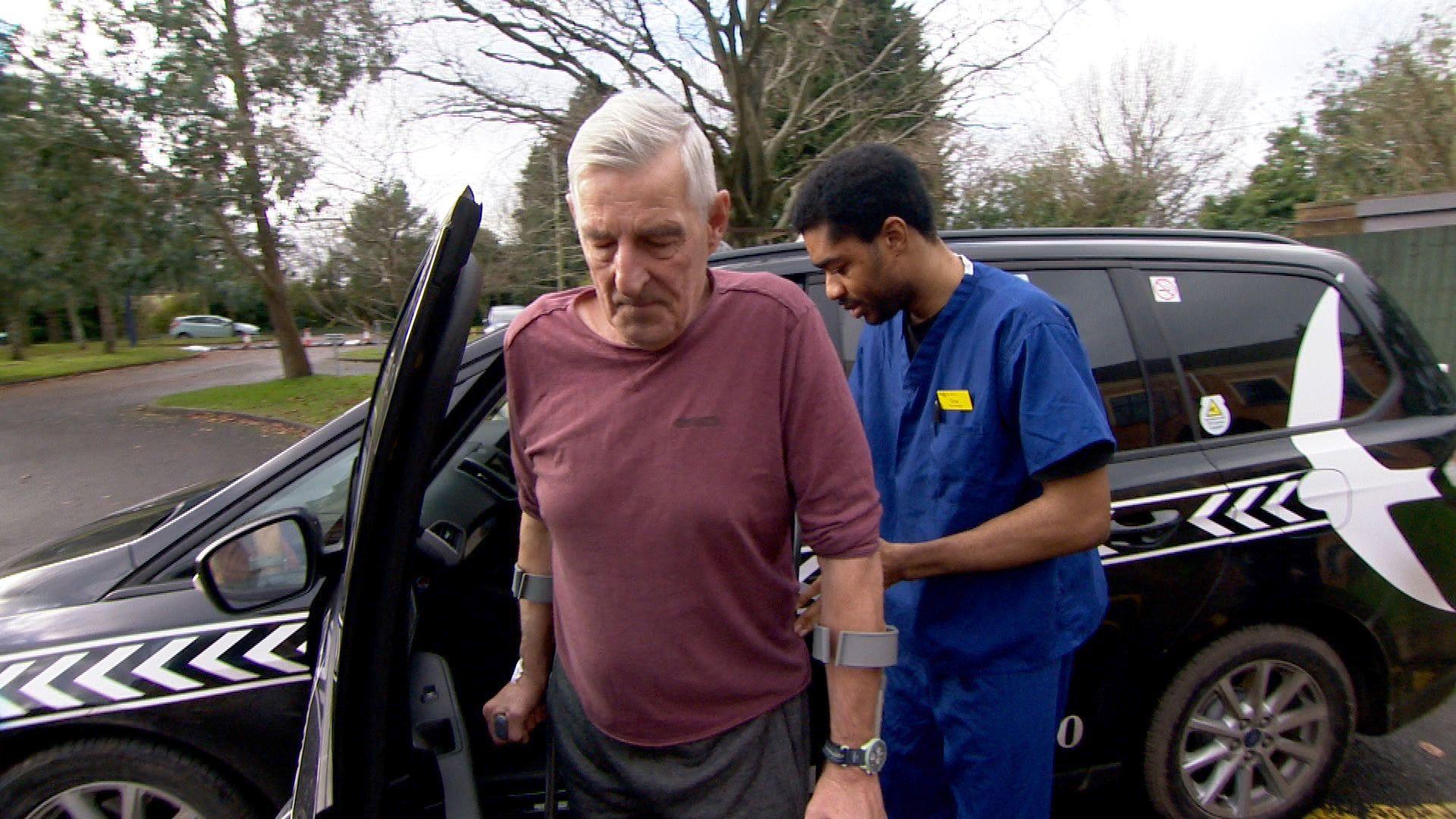 A grey-haired man using crutches being helped into a waiting taxi by a man wearing surgical clothes