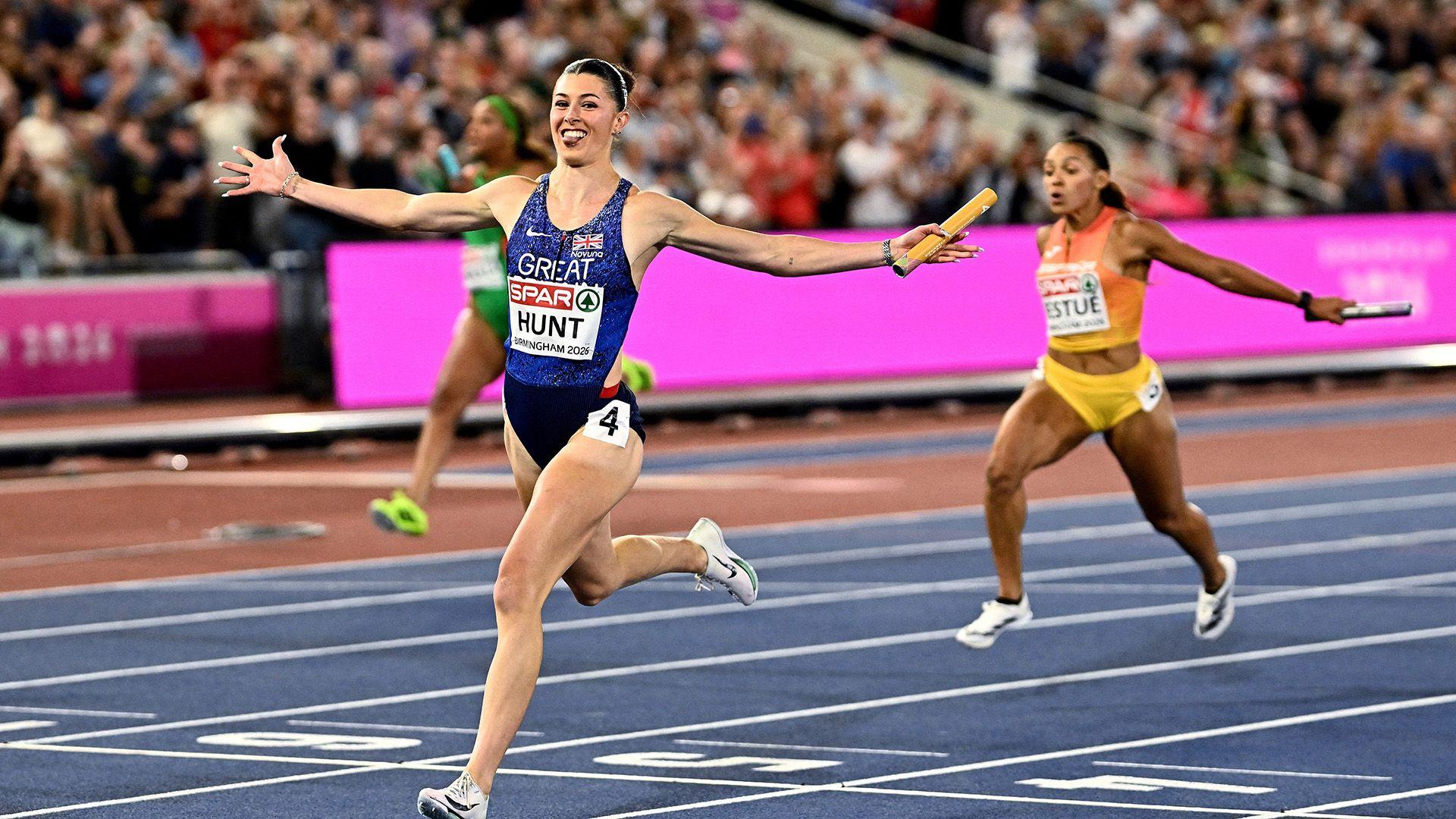 A female runner smiling holding a baton mid-race on a track with two runners behind also holding batons