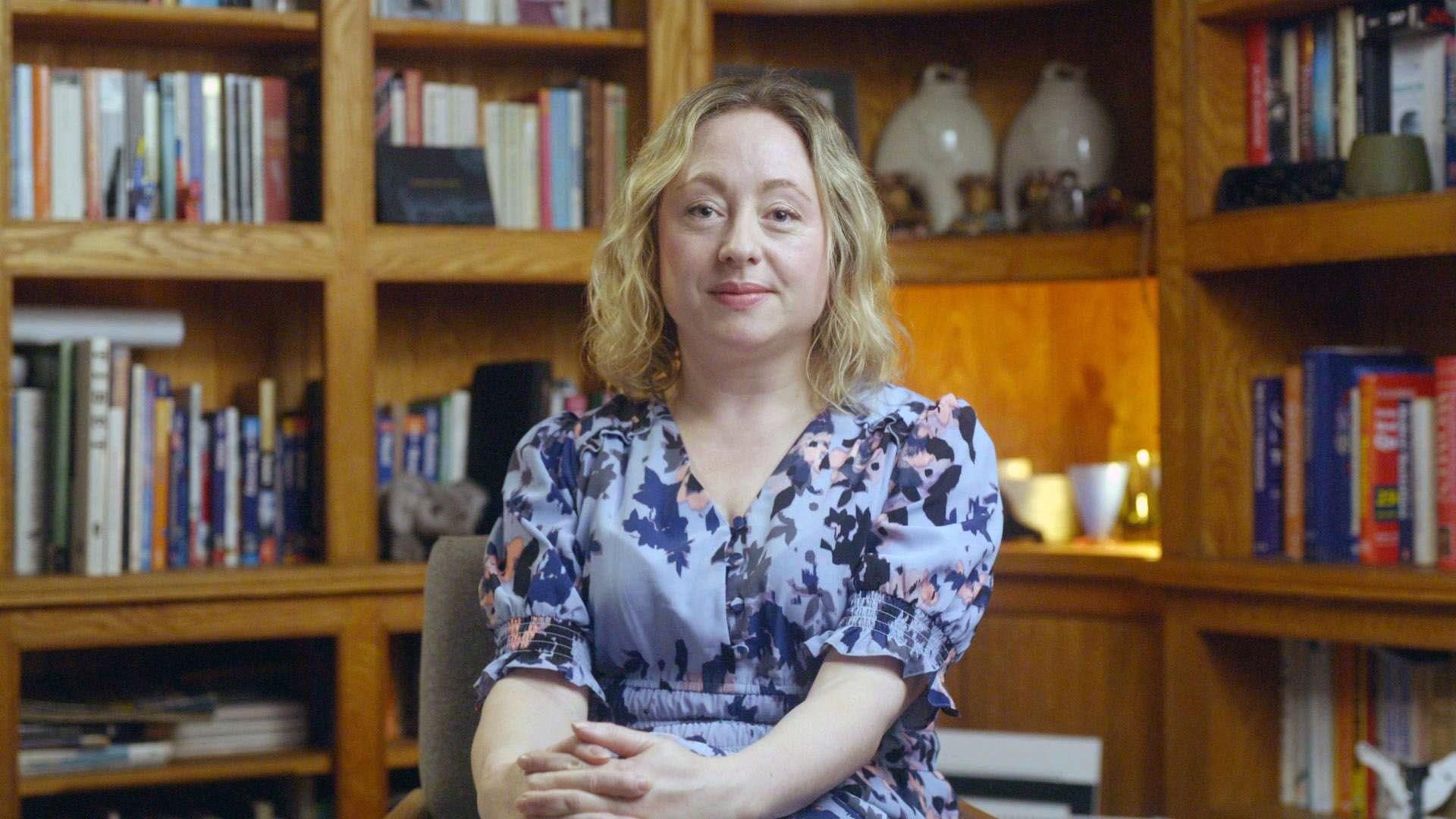 Photo of Catriona, seated, looking directly at the camera. She has shoulder-length blonde hair and is wearing a lavender-coloured pattern dress. She has her hands folded in her lap. Behind her are bookshelves containing books and other ornaments