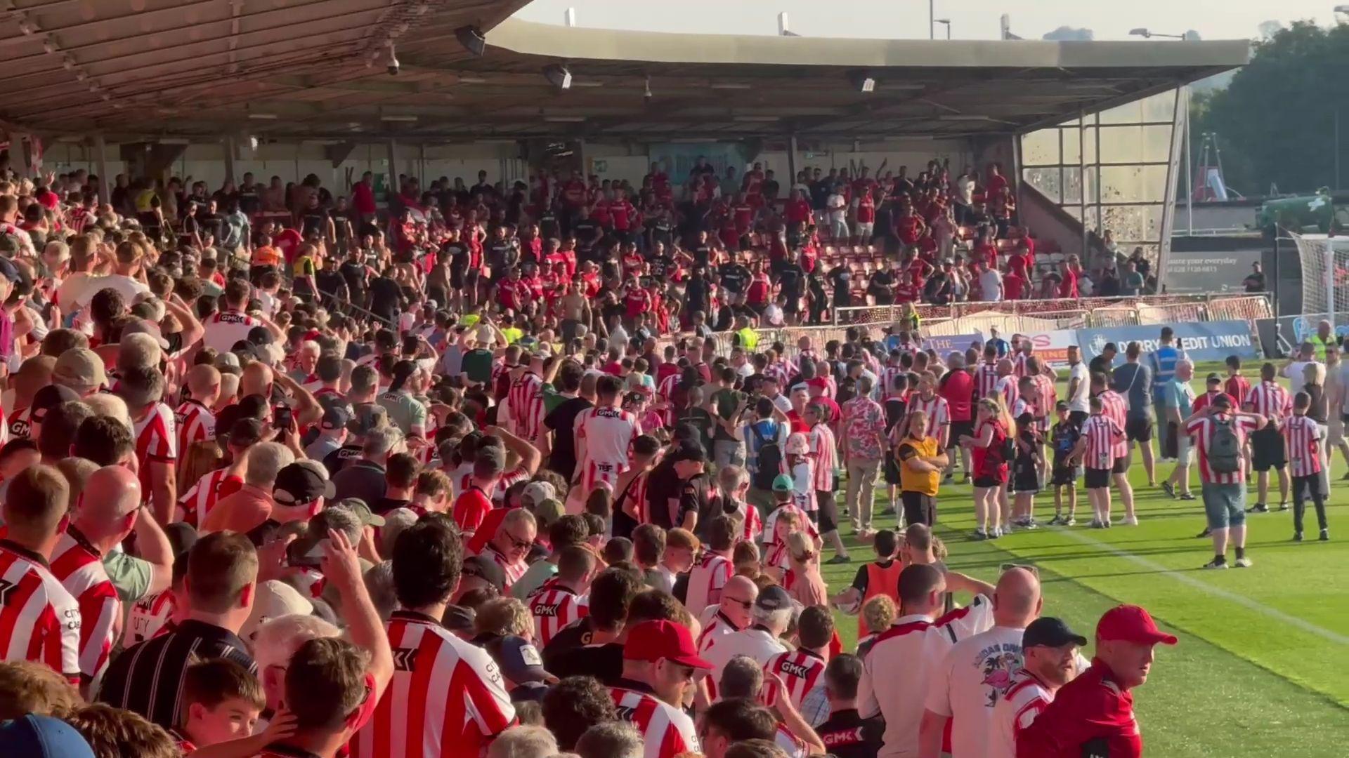 Fans in an L-shaped stand face off. Most wear red and white. Some fans are on the pitch. 