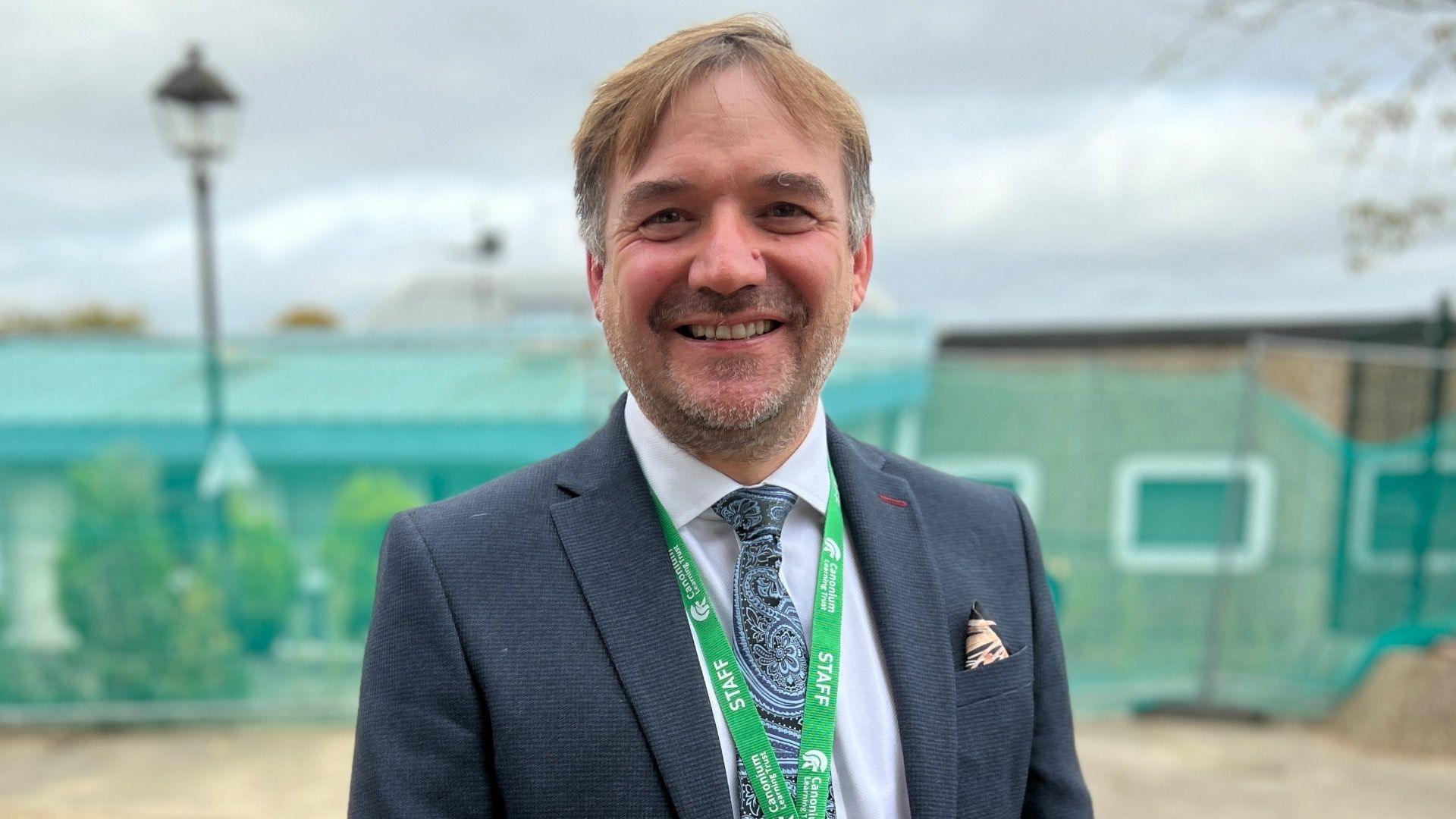 Martin Hawrylak wears a navy blue suit with a white shirt, blue patterned tie and a green lanyard around his neck. He has short brown hair and a stubbly beard, and is smiling at the camera. He stands in front of a building which has fencing around it.