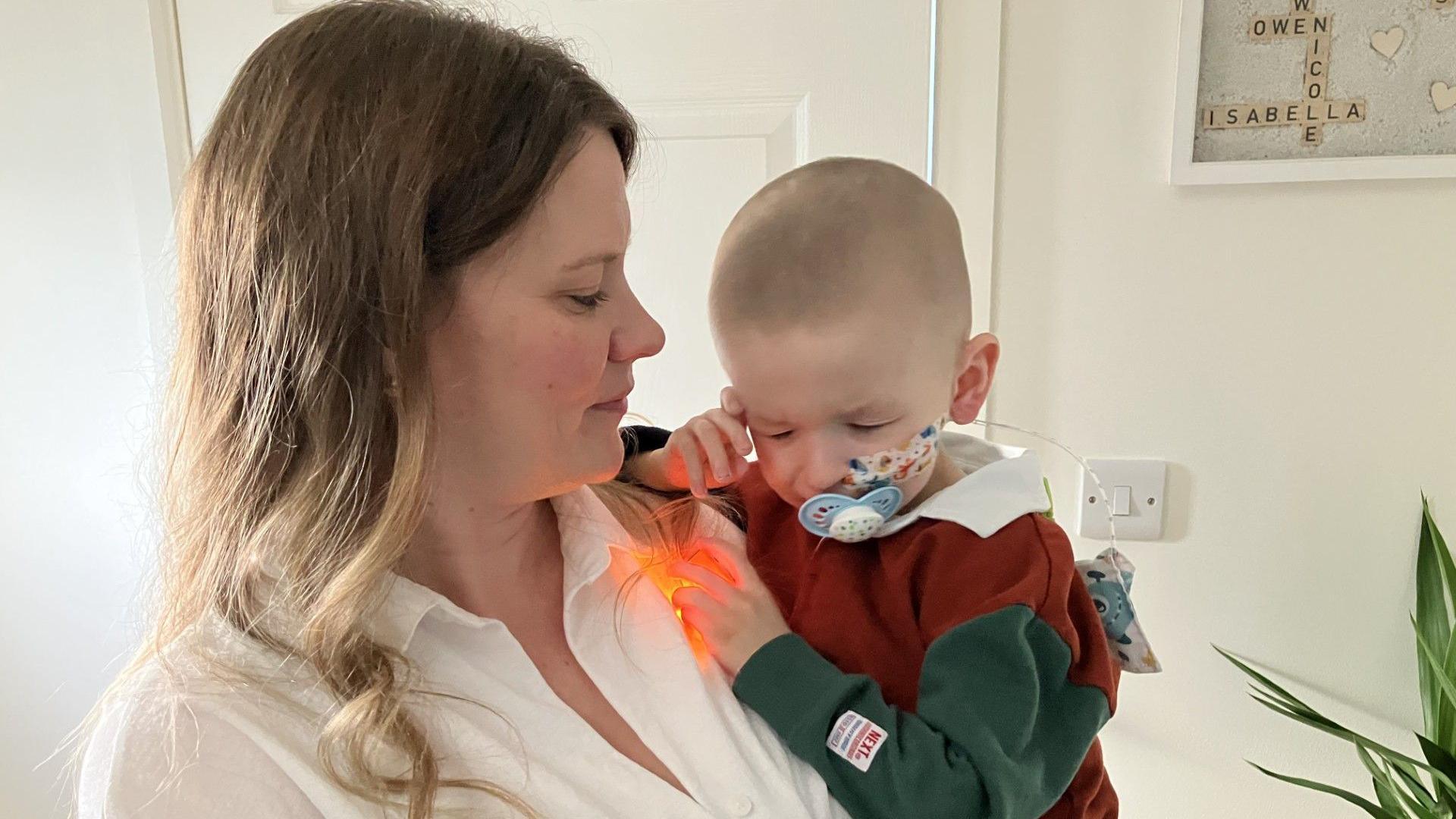 The photo shows Deborah Rowe standing and holding her son Jonah in her arms. She is looking at him and he is looking down. He has a nasogastric tube inserted and has a dummy. They are in a hallway with a cream wall behind them and a plant to the right.