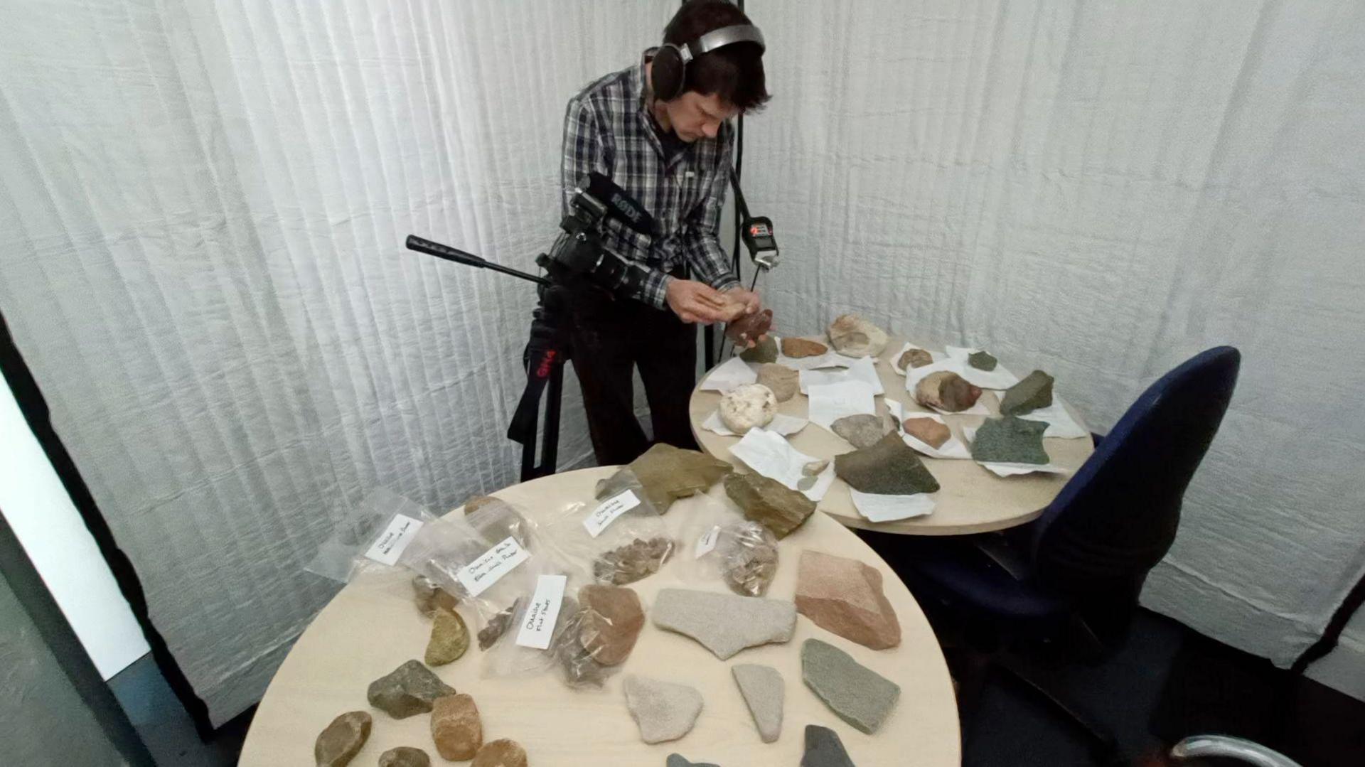 Charles Mauleverer wears headphones and is holding a microphone to a stone. He stands next to two tables which are covered with different coloured large stones.