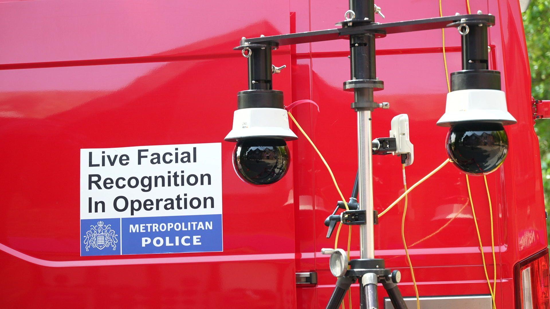 A red van, bearing a Met Police sticker reading, 'Live Facial Recognition In Operation', with two black, globe shaped cameras on a stand in front of it