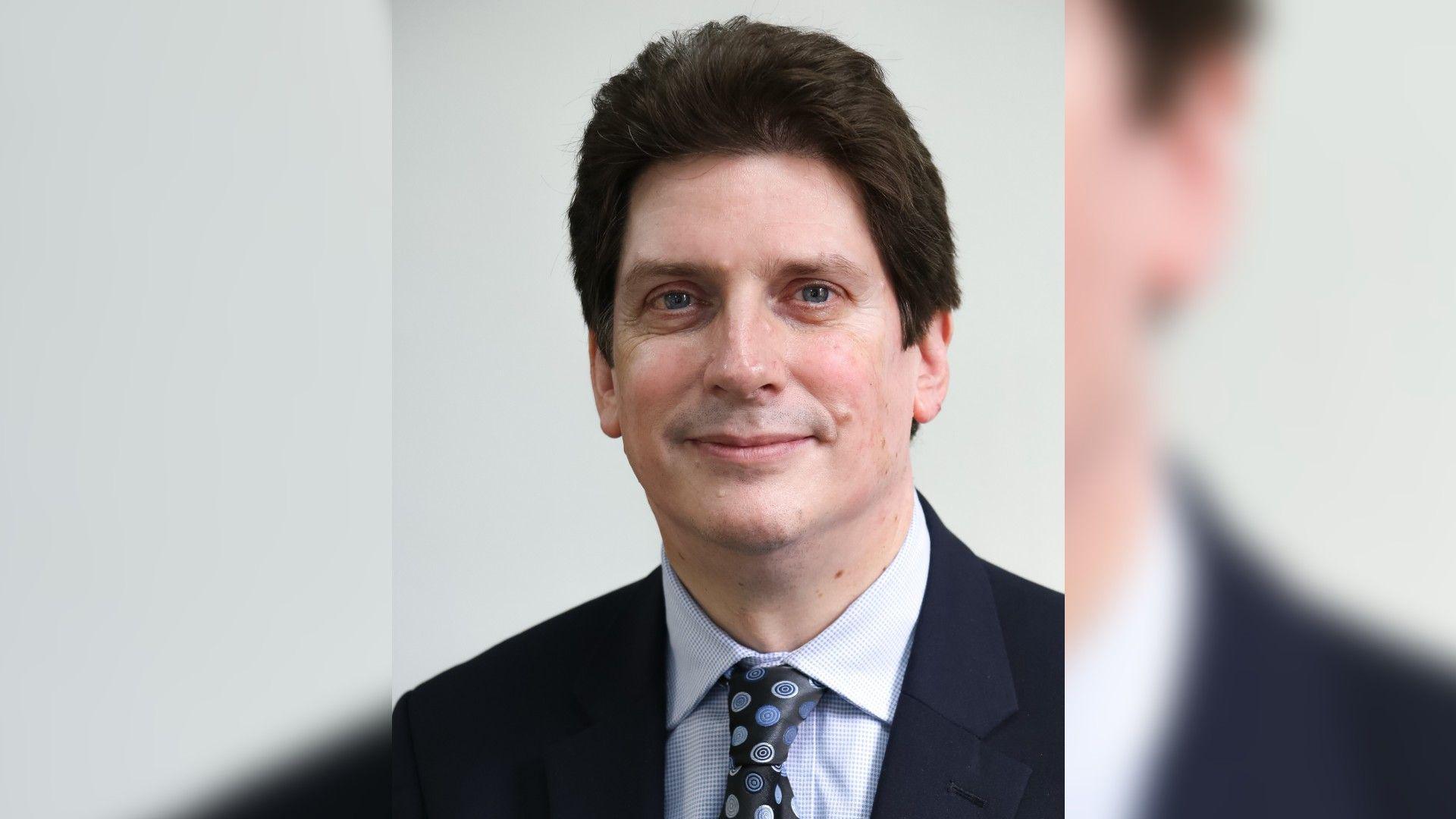 Tom Walker - a man in a black suit wearing a light blue shirt with a floral patterned black tie with blue circle flowers on it. He has dark brown hair and is smiling. White blurred background.