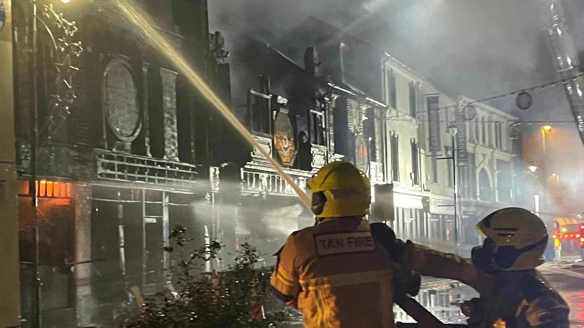 Two firefighters directing a hose at a blackened building 