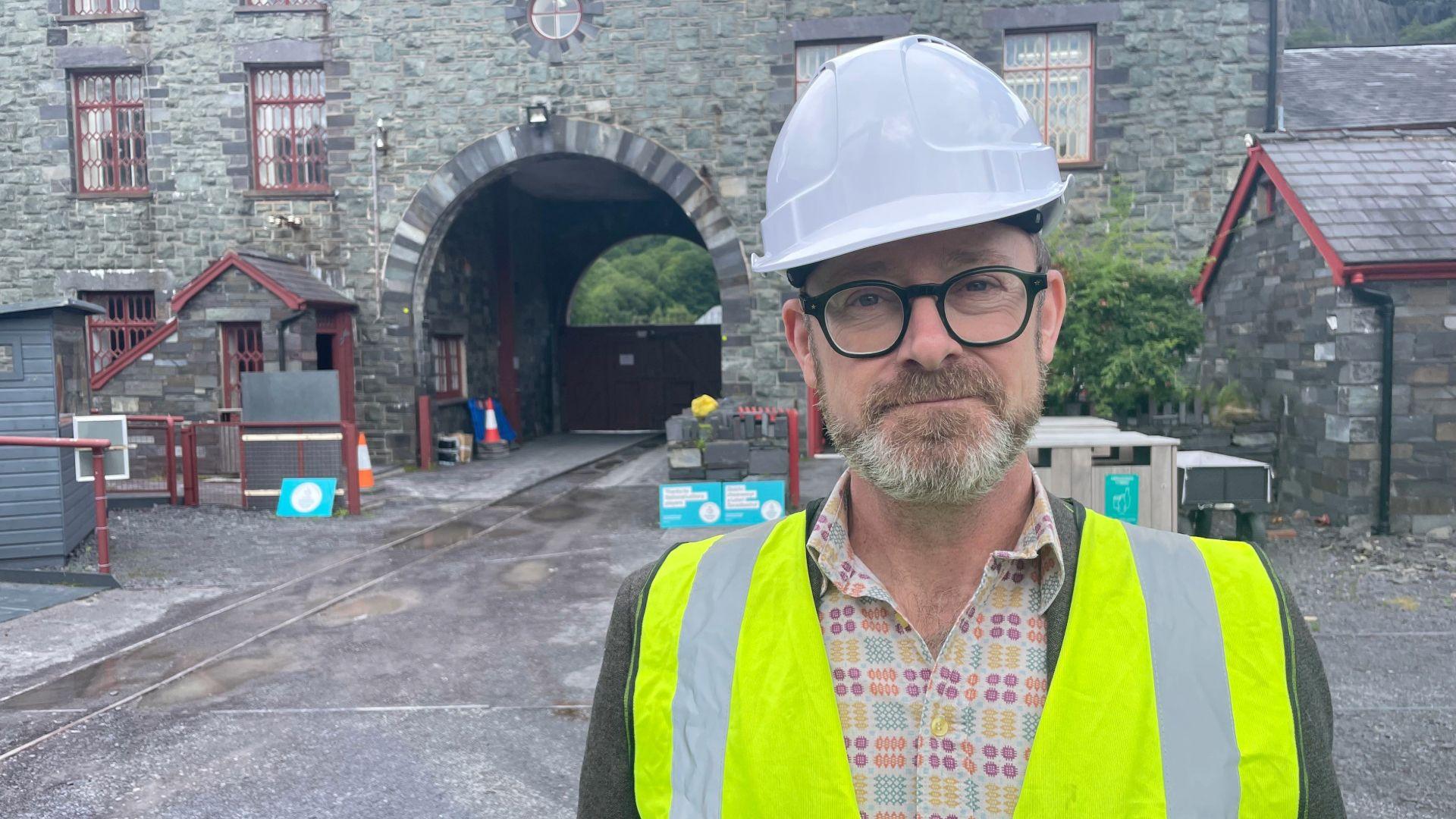 Llun o Andrew White yn sefyll ar safle'r amgueddfa. Maen gwisgo helmed gwyn a fest 'high-vis'. Adeilad cerrig yn y cefndir a 