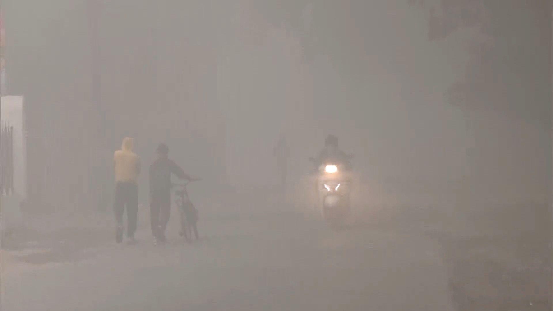 A couple is seen walking with a bicycle while a man on a scooter passes them in dense fog