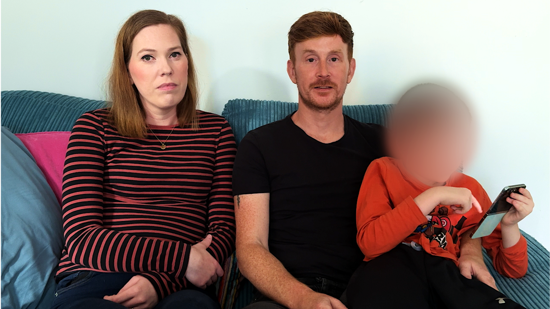 A family of three sit on the sofa. The woman has red hair and is wearing a red striped top. Next to her is a red-haired man in black, and on his lap is a boy in orange whose face is blurred.