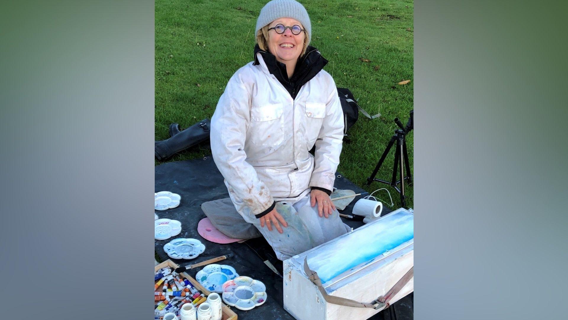 A woman in a light-coloured protective suit and woolly hat kneeling on a mat outdoors beside a selection of watercolour paints, brushes and a painting box on grass.