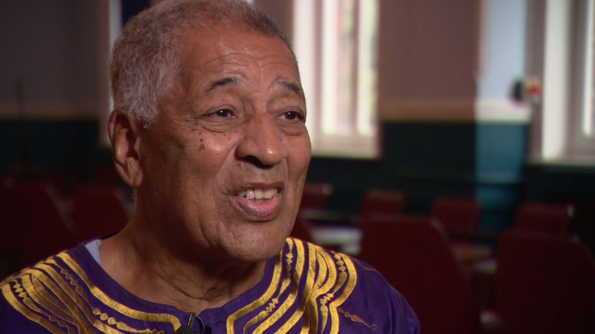 A man wearing a purple African top with gold brocade smiles into the camera