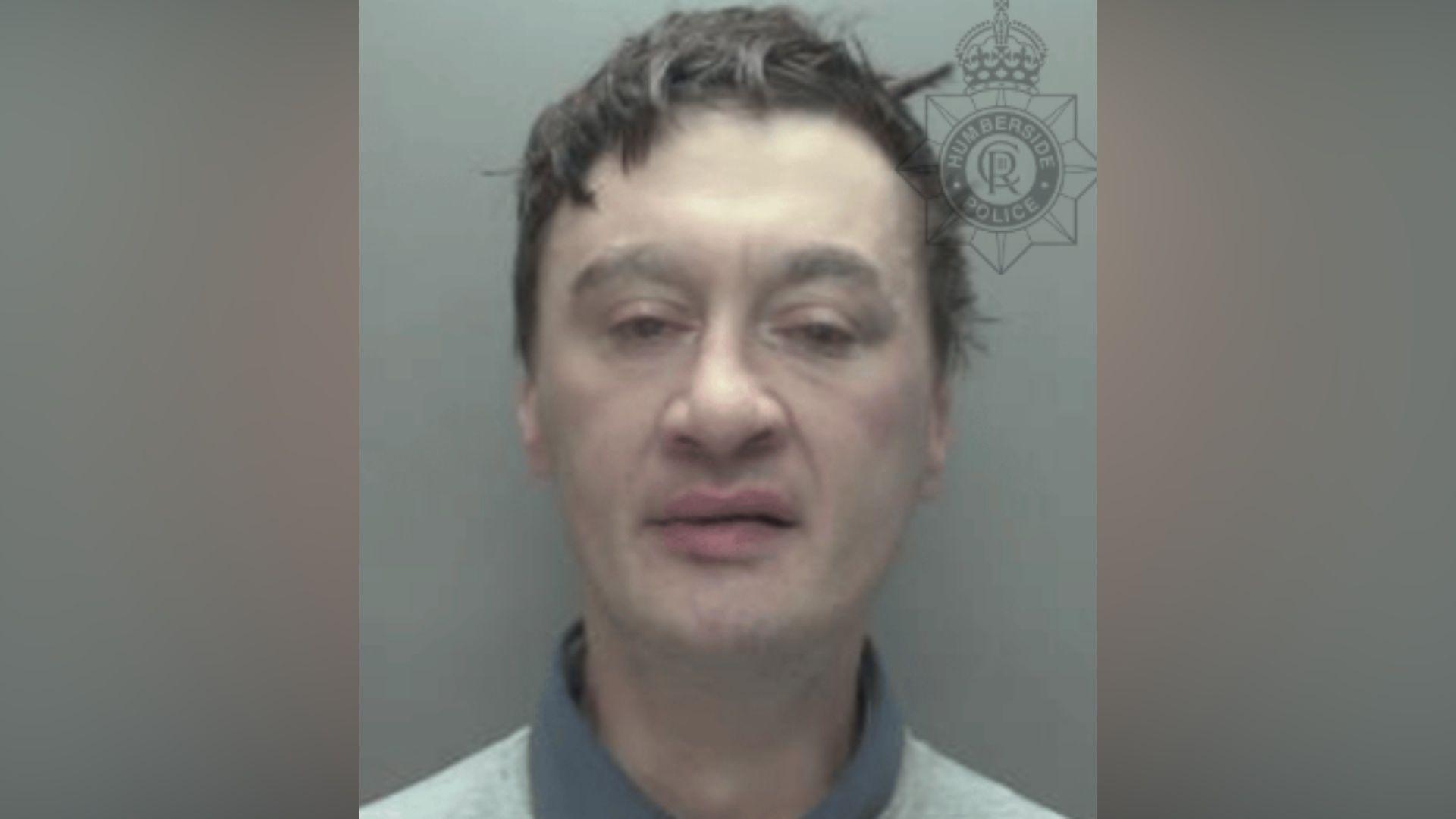 A man with messy short black hair in a police mugshot. His eyes are half-closed.