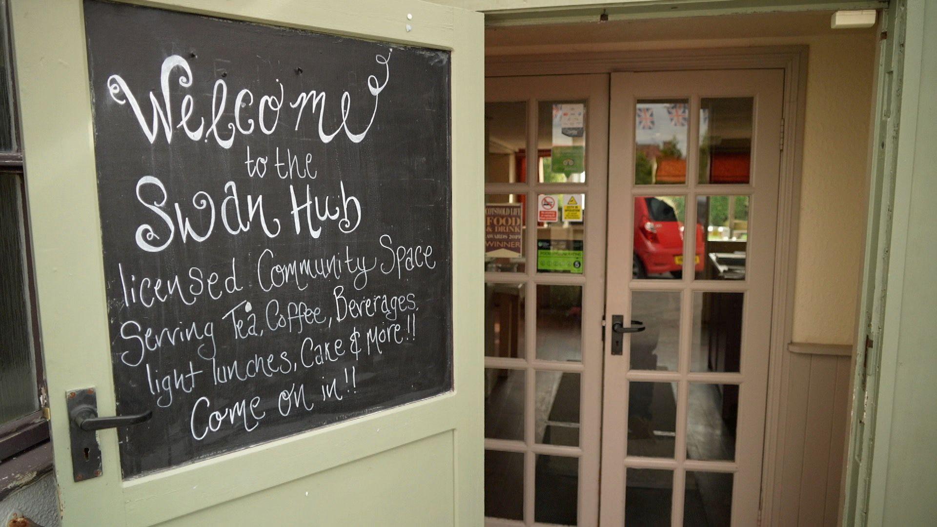 A view into the entrance of the Staunton and Corse Community Hub, through the sage-green exterior door of the Swan pub. There is a chalkboard on the inside of the door with scrawled white handwriting welcoming visitors inside. 