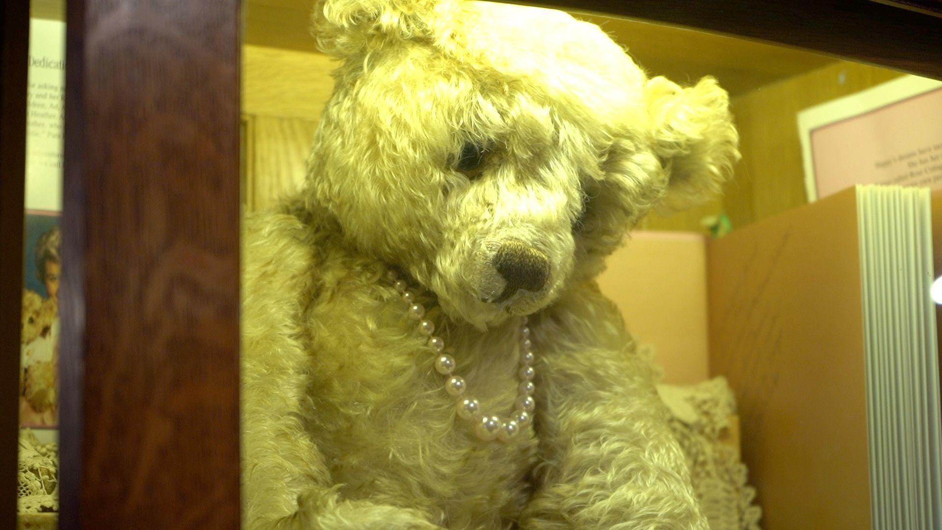 A teddy bear with a light fur and a pearl necklace displayed behind a glass cabinet. There are books behind it.