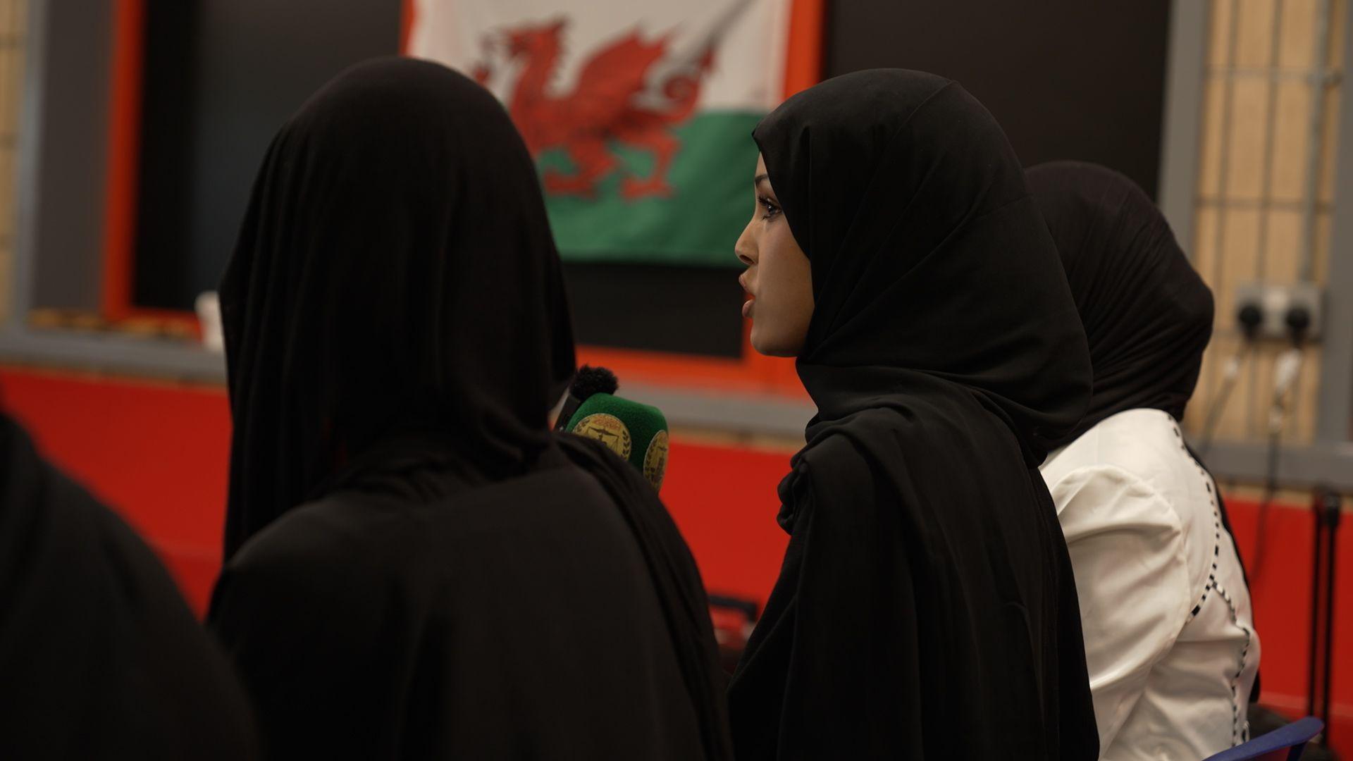 A young Somali woman in a black head scarf and Abaya speaks into a microphone alongside her peers, her face in profile. A Welsh flag is visible in the background.