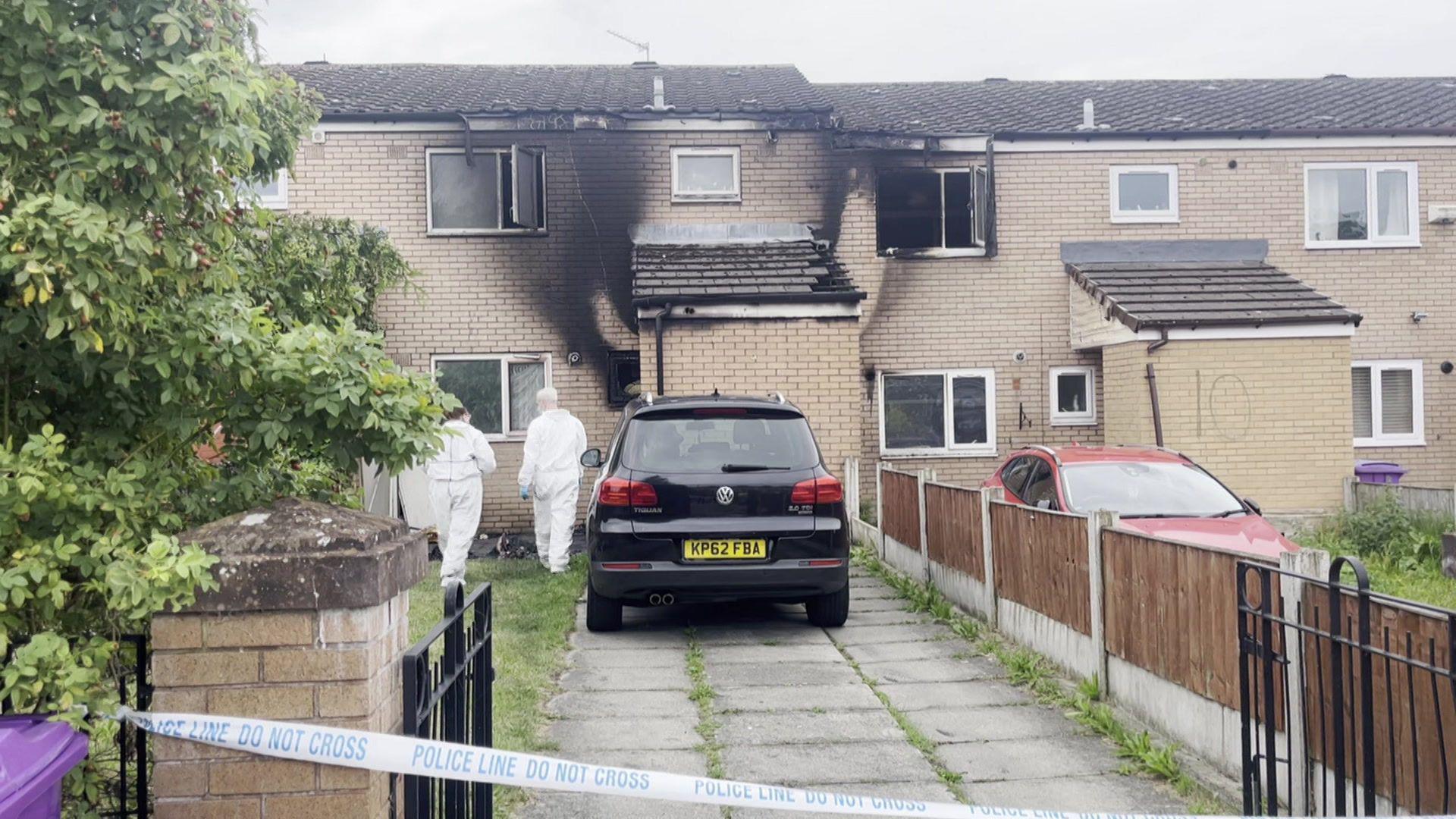 Two terraced houses with visible smoke damage on the brickwork and windows. Two forensic investigators in white overalls approach the front door of the house on the left, which is cordoned off with blue and white police tape. 