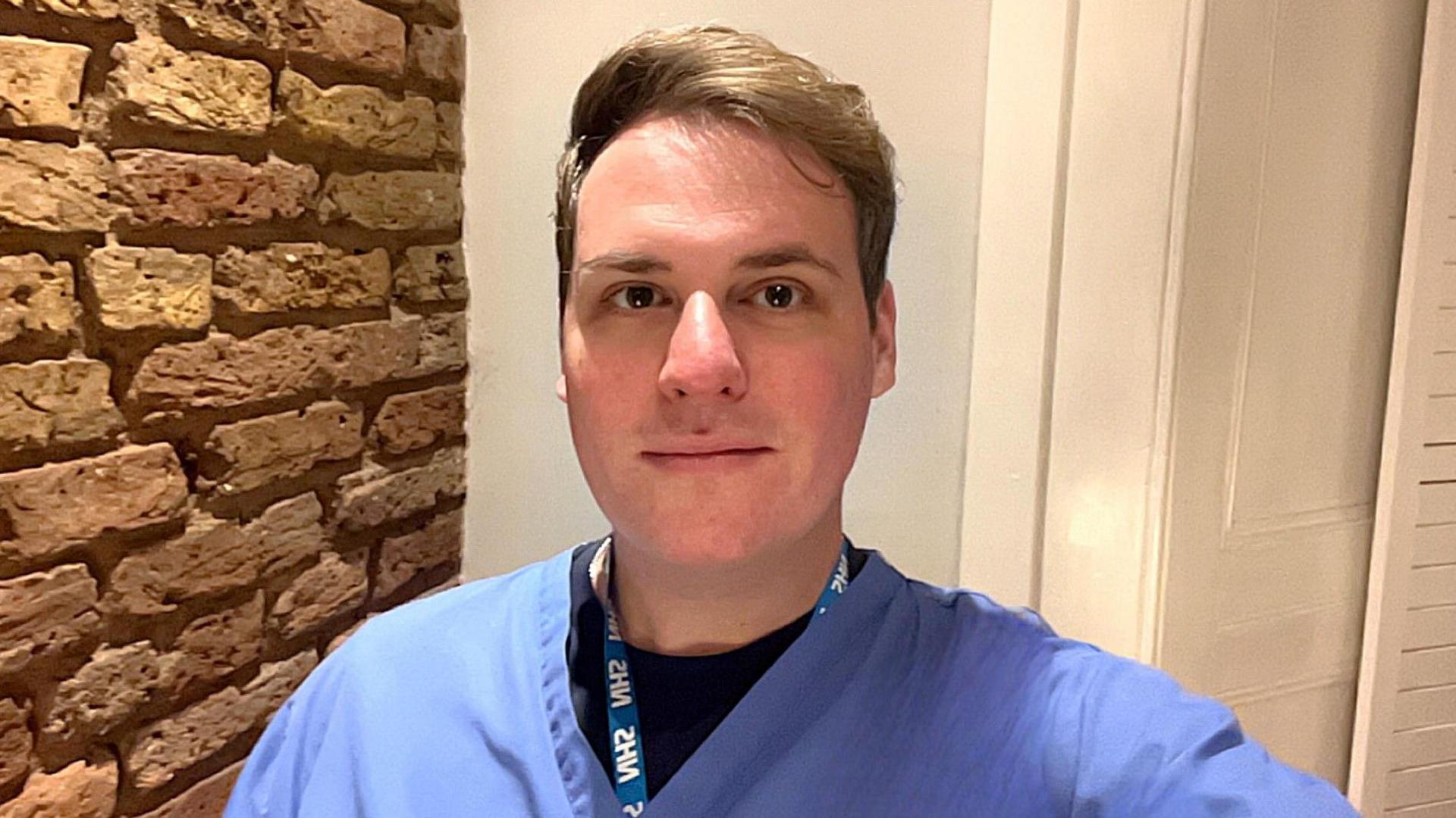 John Watt wearing his blue dentist scrubs and an NHS lanyard. He has short brown hair swept over to one side and is smiling at the camera, standing beside a brick wall