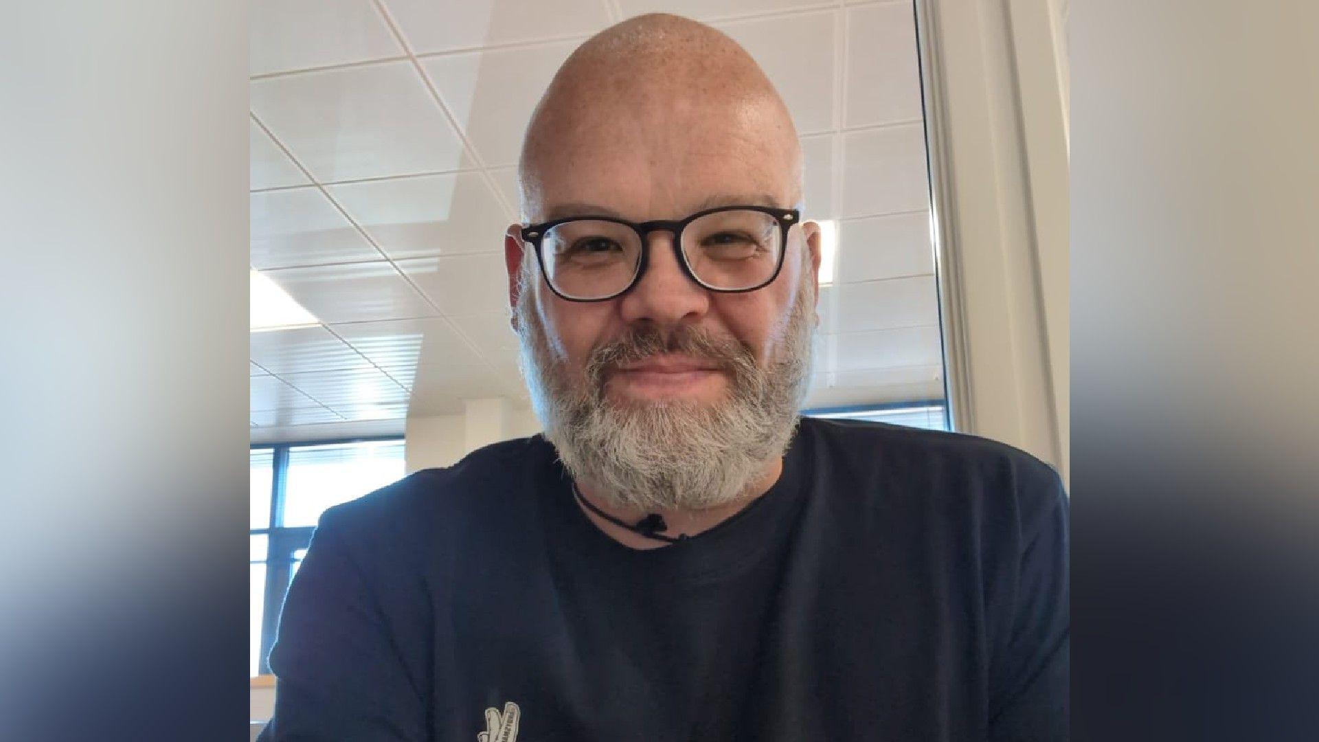 A selfie of a bearded and bespectacled man who smiles at the camera. He is wearing a blue top with a white logo on his chest.