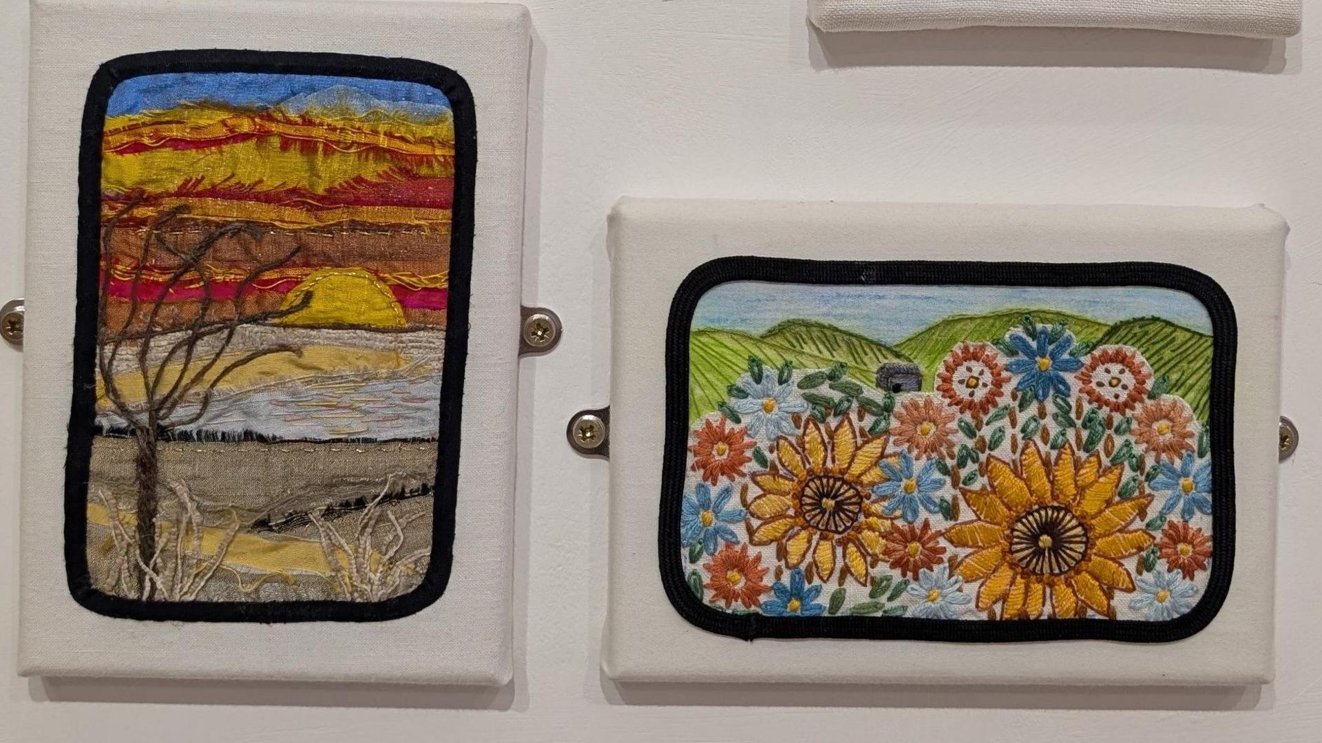 Two pieces of embroidery have been placed on a white wall. One shows a sunset with a bare tree in the foreground and the other shows two sunflowers with green fields in the background.
