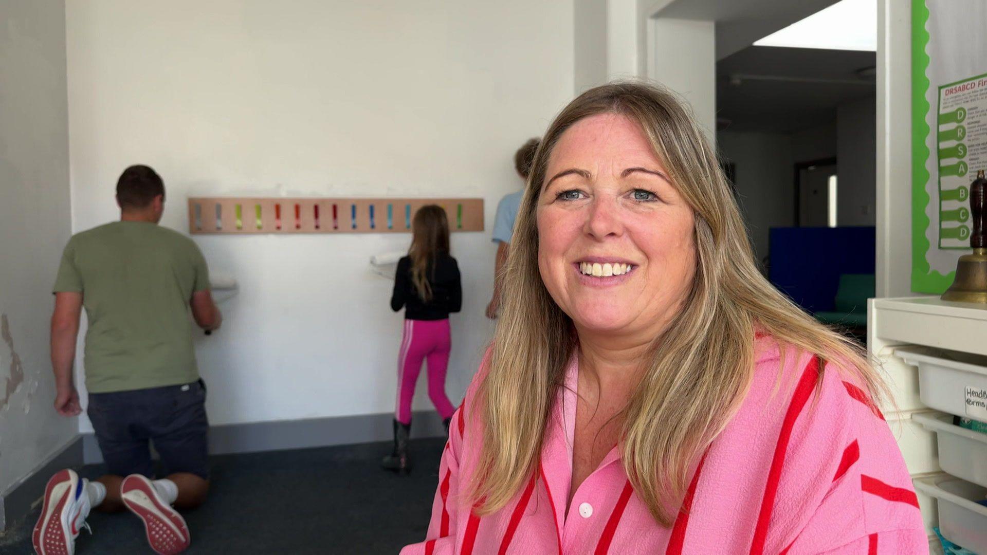 Louise Shore sits and smiles for the camera. She is wearing a large pink short with red stripes and has long straight blond hair. In the background a man and child can be seen painting a while wall.