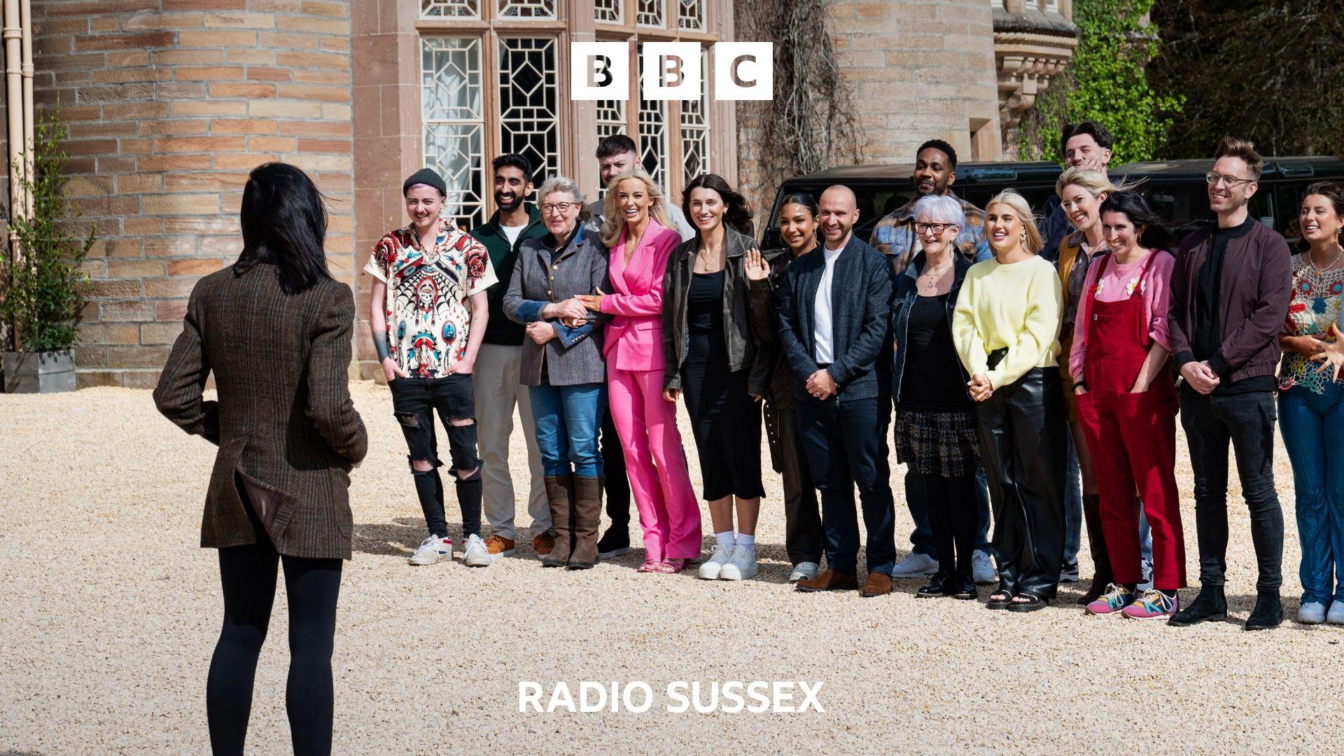 The contestants from series 3 of The traitors are lined up outside Traitors Castle being greeted by Claudia Winkleman. Claudia has her back to the camera and her hands in her pockets. She is wearing a brown jacket and black trousers. The contestants are dressed in various outfits and stand, smiling nervously.