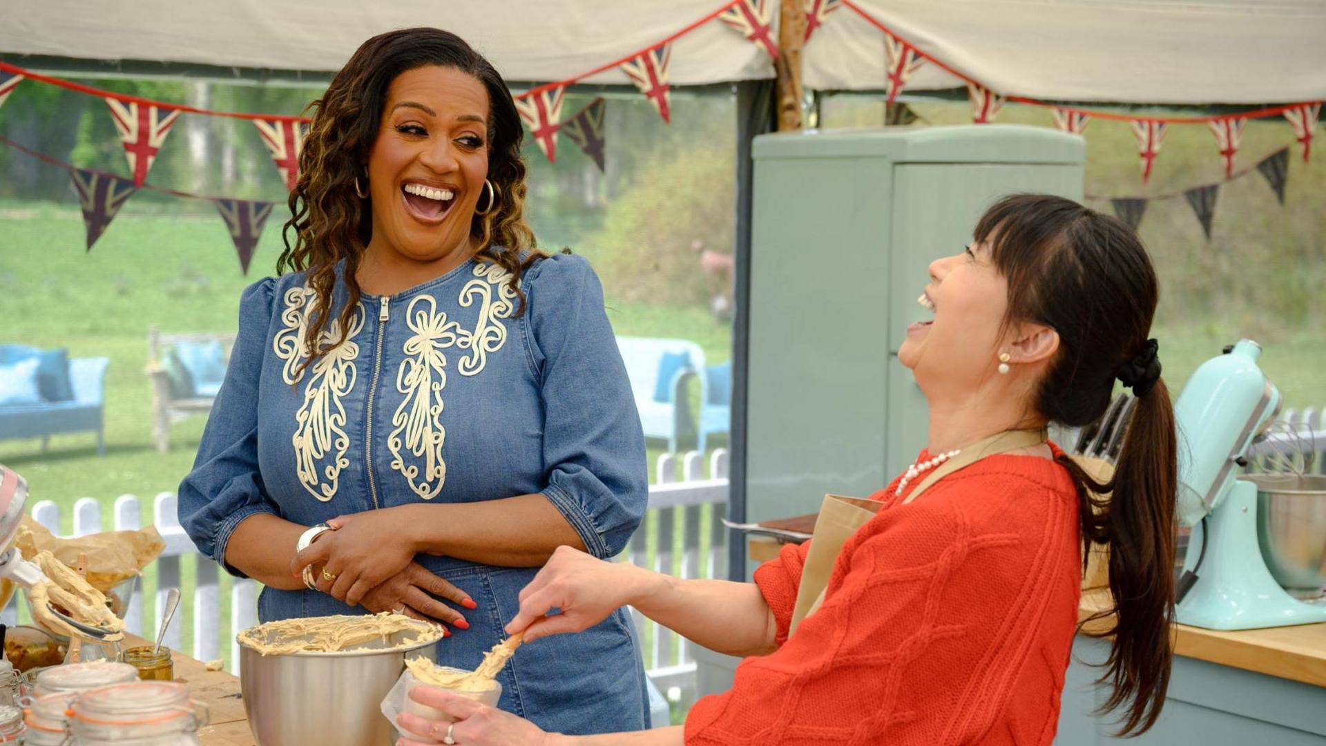 Pui Man throws her head back in laughter while putting cake mixture into a silver bowl. She has her hair in a ponytail and is wearing a red jumper. In front of her is TV presenter Alison Hammond, who is wearing a blue dress and is also laughing.