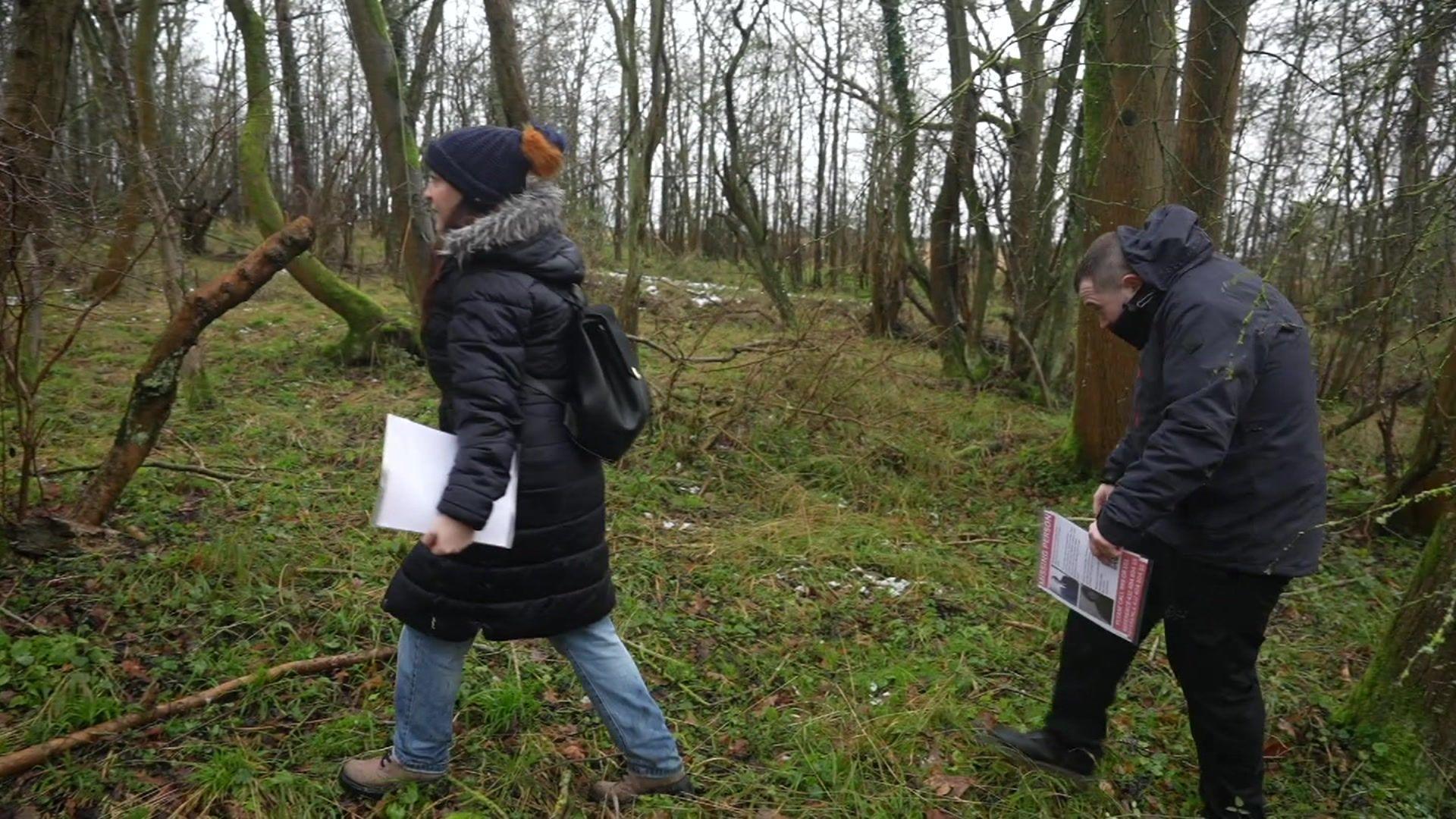 A woman and man walk through woodland wearing outdoor winter clothing and carrying posters about missing Thomas Roche.