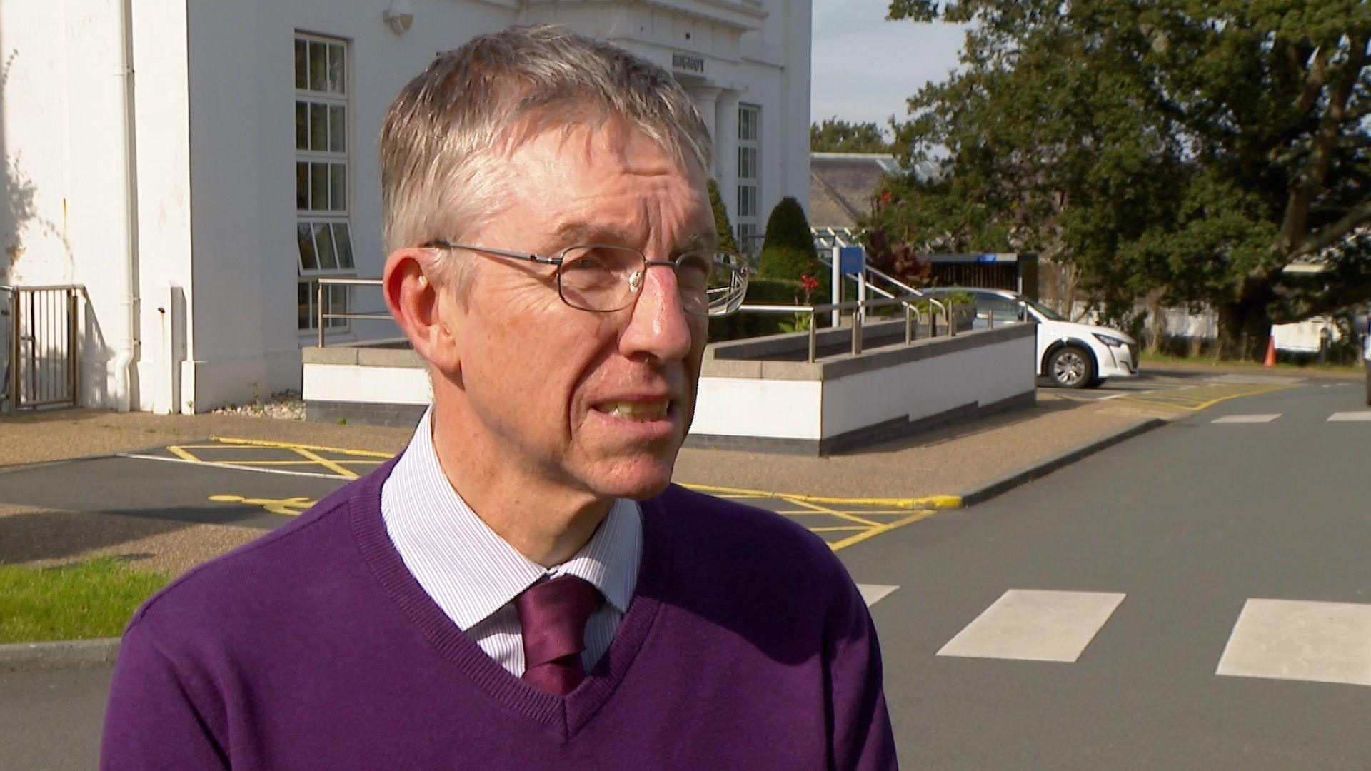 A man wearing a purple jumper with a white shirt, a purple tie and glasses.