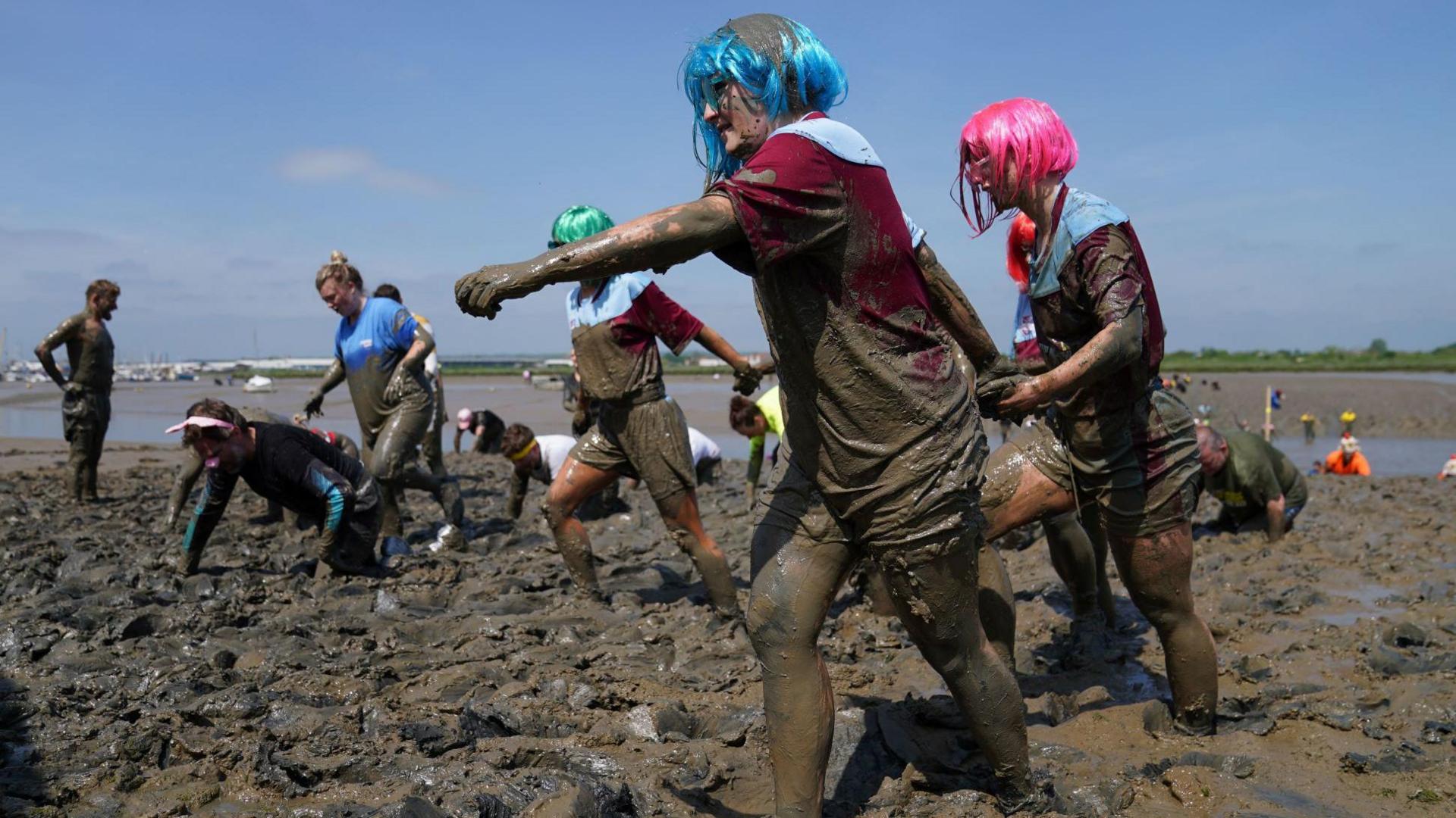 People walking and crawling through the thick mud during a previous race. In the foreground, two people wearing colorful wigs march through the mud while holding hands.