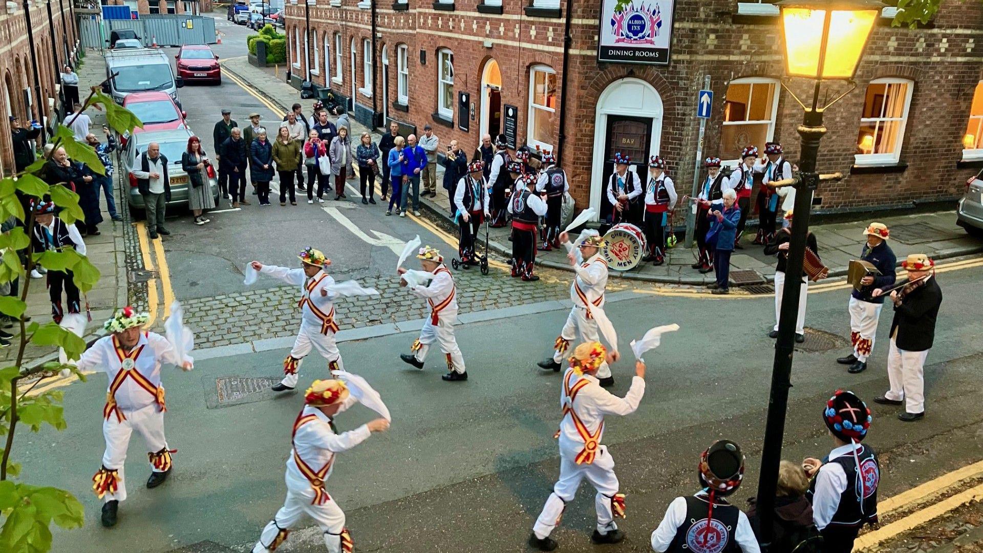 A wide angle shot outside the Albion Inn. Performers are dancing in the street with an audience from the local neighbourhood watching.