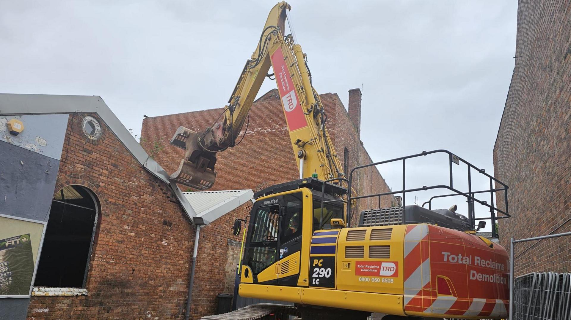 A bright yellow demolition vehicle tears the corrugated metal roof from the top of the building.