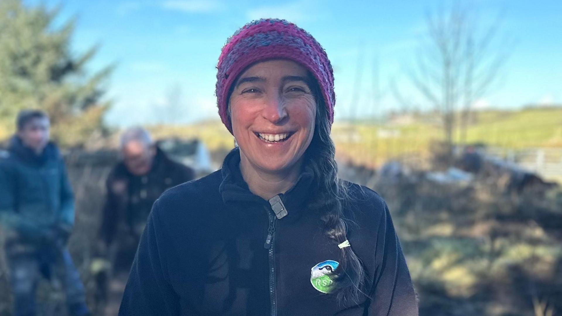 Anne smiling at the camera as she stands in a wooded area. She is wearing a black zip-up fleece with an RSPB badge, a pink and blue hat, and has her dark brown hair in a plait.