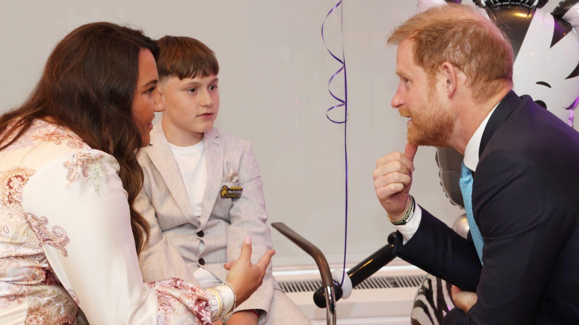 Prince Harry sits on the right wearing a dark blue suit, with a white shirt and sky blue coloured tie. He is talking to Jude, who is 14 and has brown short hair. Jude is wearing a beige linen two-piece suit which has dark brown buttons on the blazer and has a white cotton t shirt underneath. He is sat in a chair looking at Prince Harry, who is talking to a woman with dark brown hair, wearing a paisley printed silk blouse sat next to Jude. There is a helium inflated zebra balloon in the background.