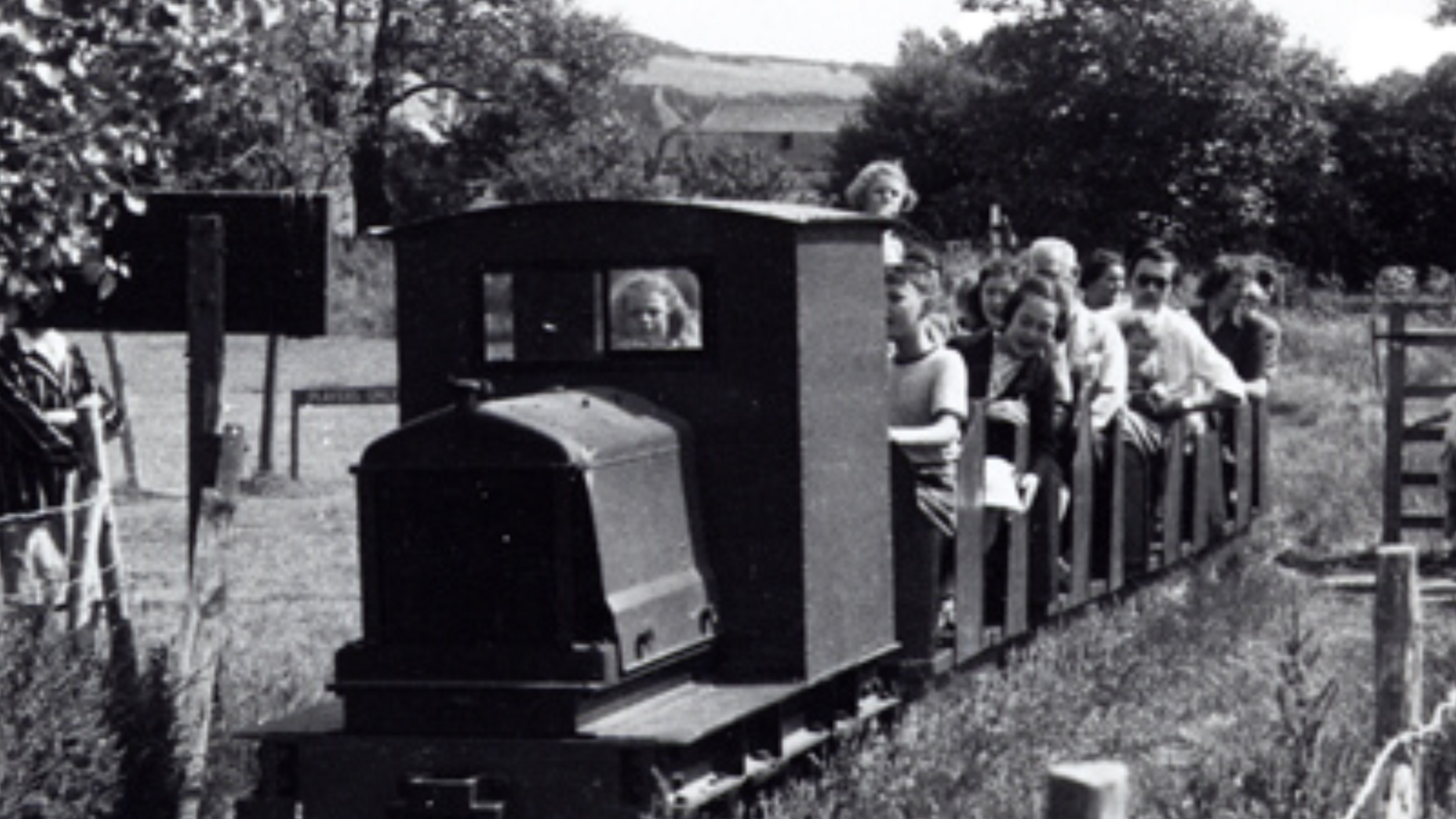 A black and white image of the Drusillas miniature train. The train has a group of people sat in rows behind it.