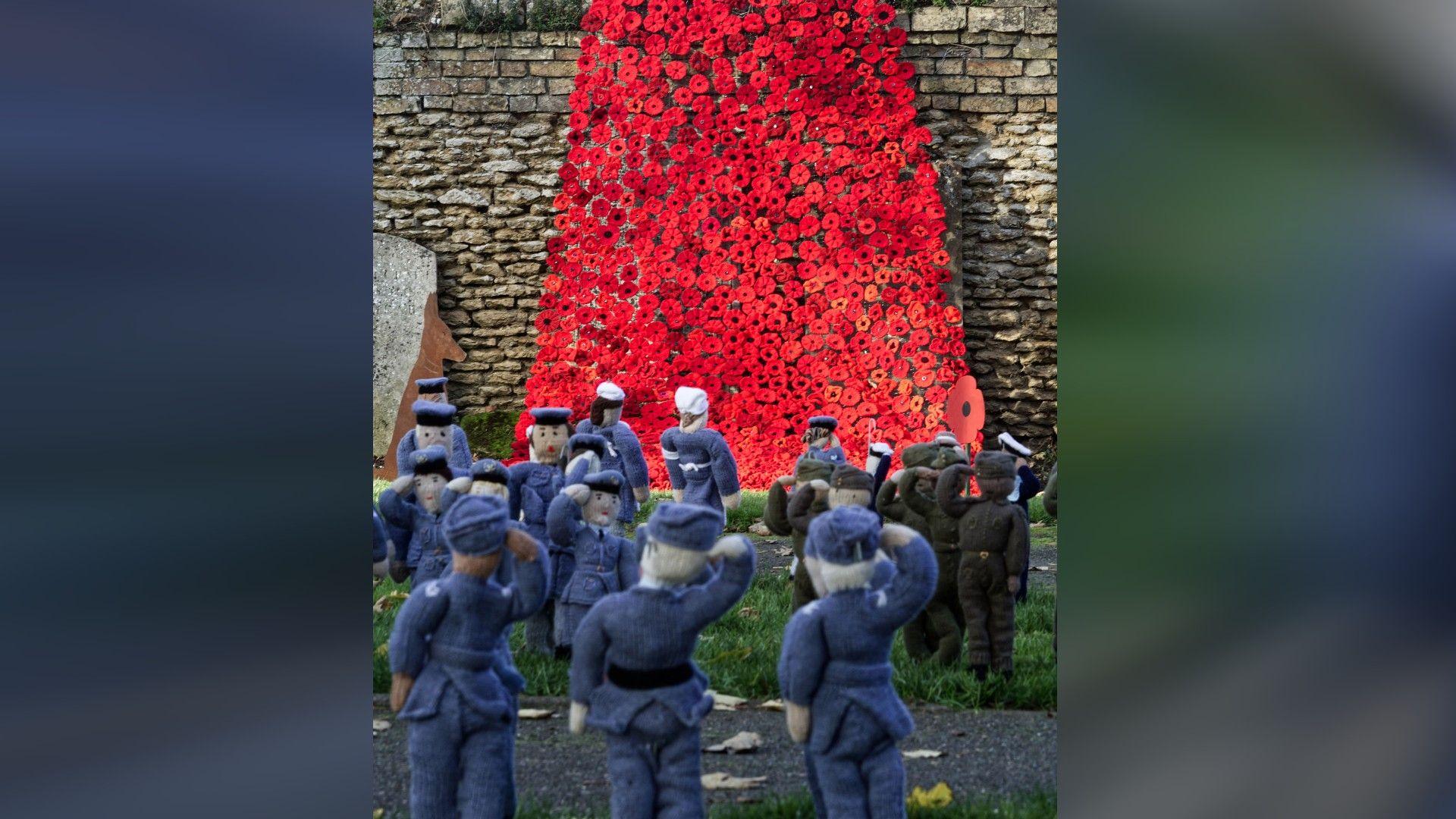 A waterfall of red poppies on a stone wall with small knitted soldiers on the grass in front of it 