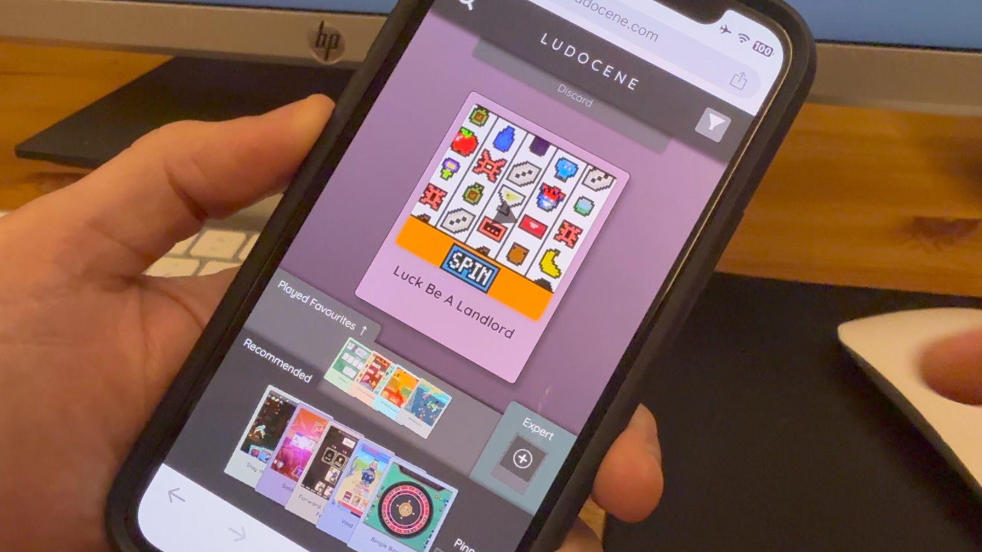 A close-up of a mobile phone in someone's hand. The screen is arranged into rows of cards. The top displays a single central card showing a screenshot from Luck Be A Landlord. Below that are two rows titled "played favourites" and "Recommended" containing multiple cards showing screenshots and titles from different games. 