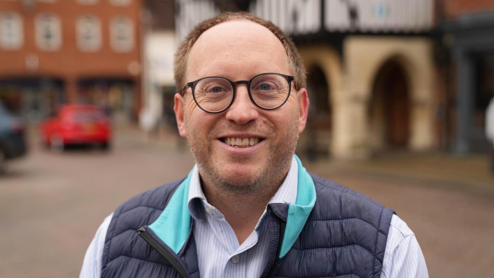 Conservative association chairman George Smith smiling at the camera in the Saffron Walden. He has receding brown hair and a beard. He is wearing a blue gilet and a blue-and-white striped shirt.  