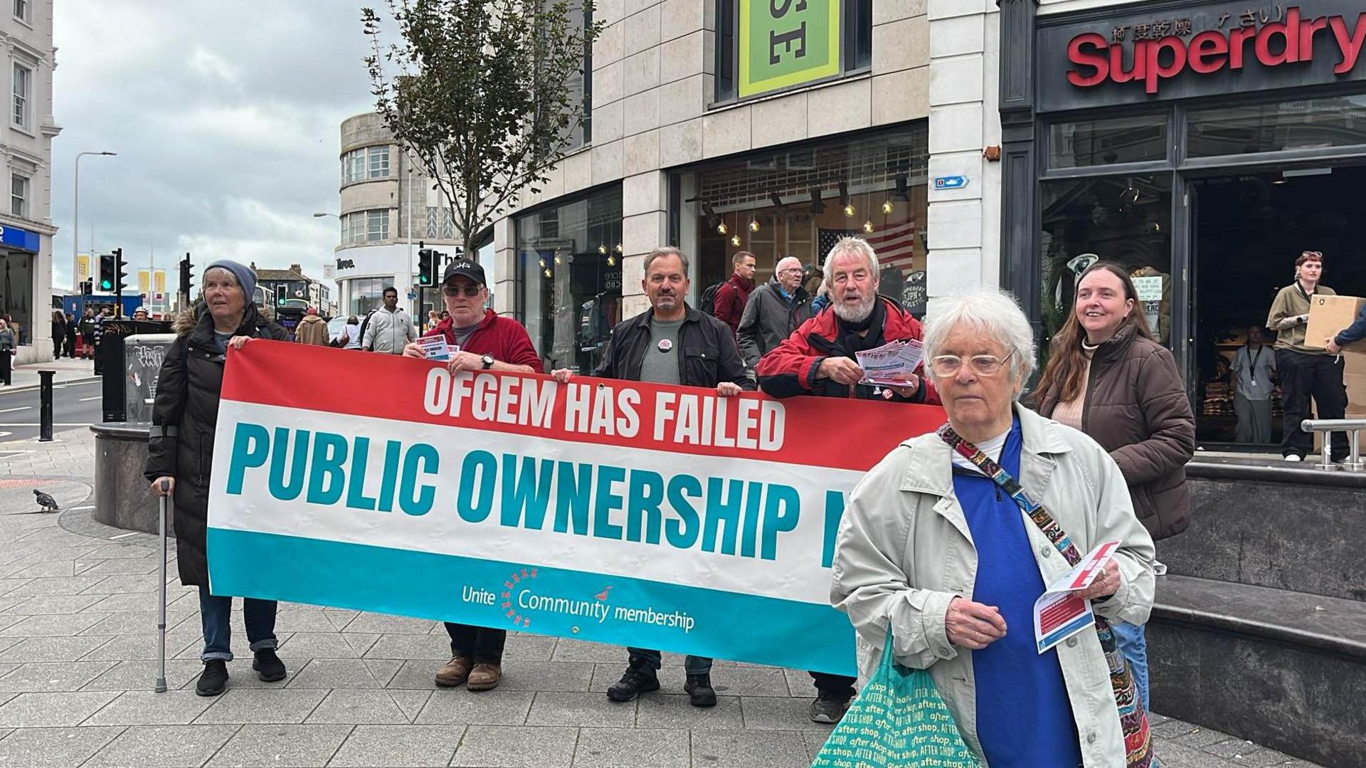 Five Unite members in the centre of Brighton, campaigning against the increase in the energy price cap, holding a banner, as a female pensioner walks away.