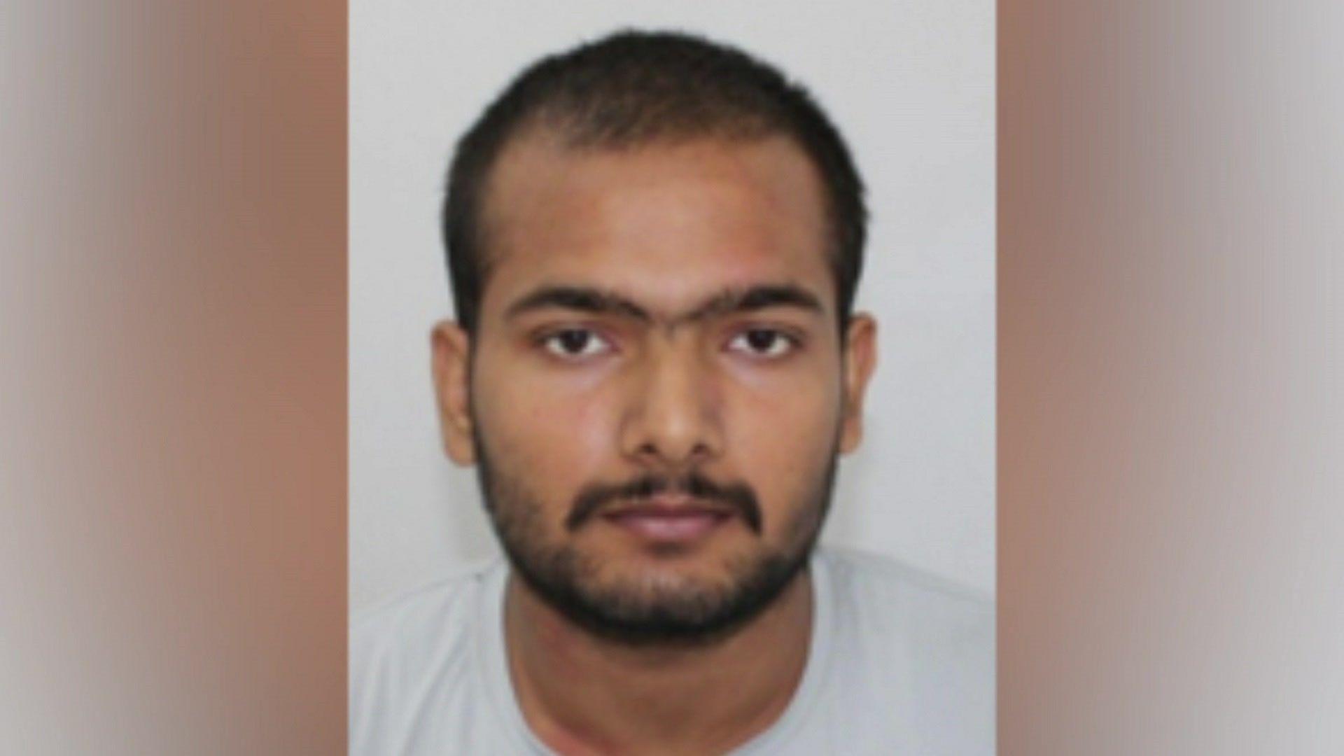 A police mug shot of an man with a short haircut and a beard wearing a grey t-shirt looking directly at the camera