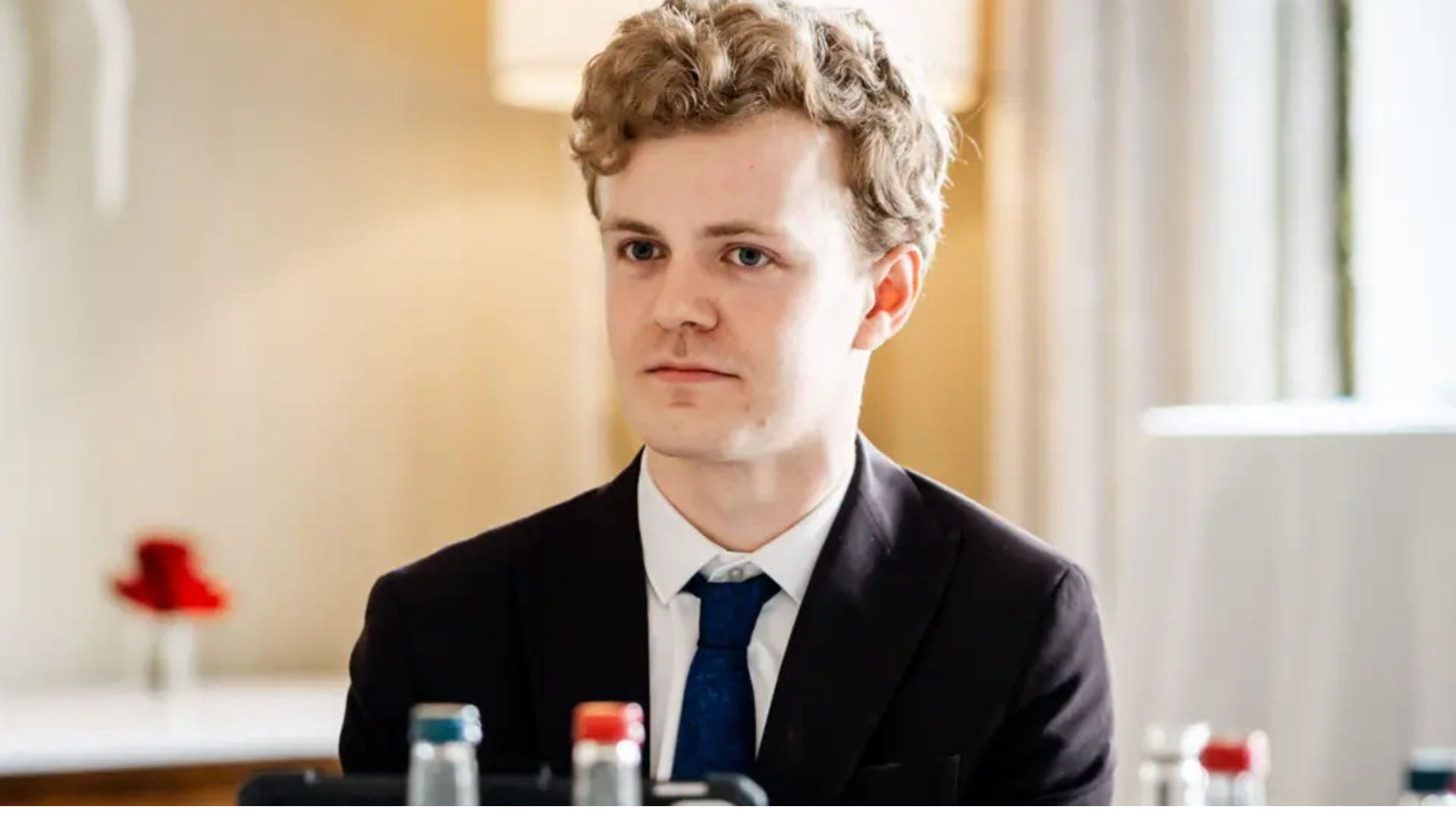 Sam Carling wearing a black suit with a blue tie and white shirt. He has curly strawberry blond hair, is clean shaven, and is sitting in a well-lit room.