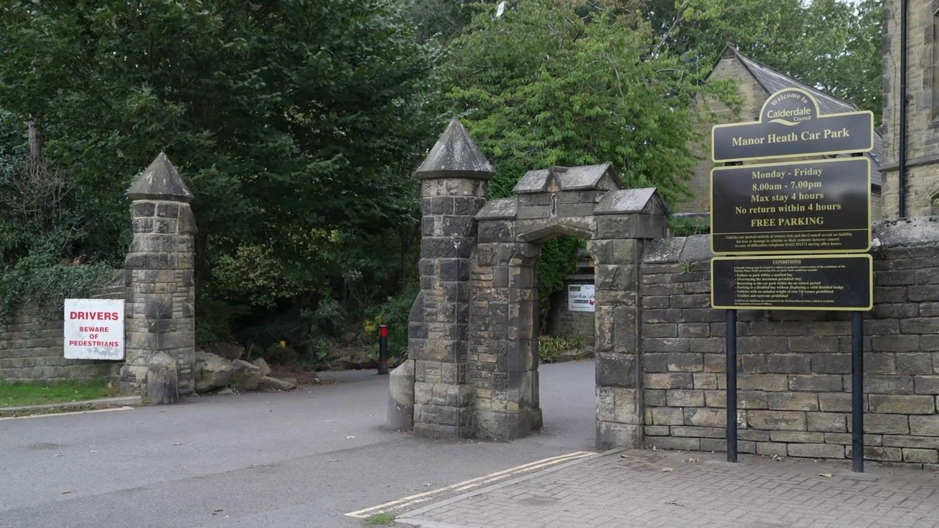 A grand entrance to a park with a sign to the right that reads: Manor Heath Car Park. There are trees and stone buildings and a road leading between stone pillars.