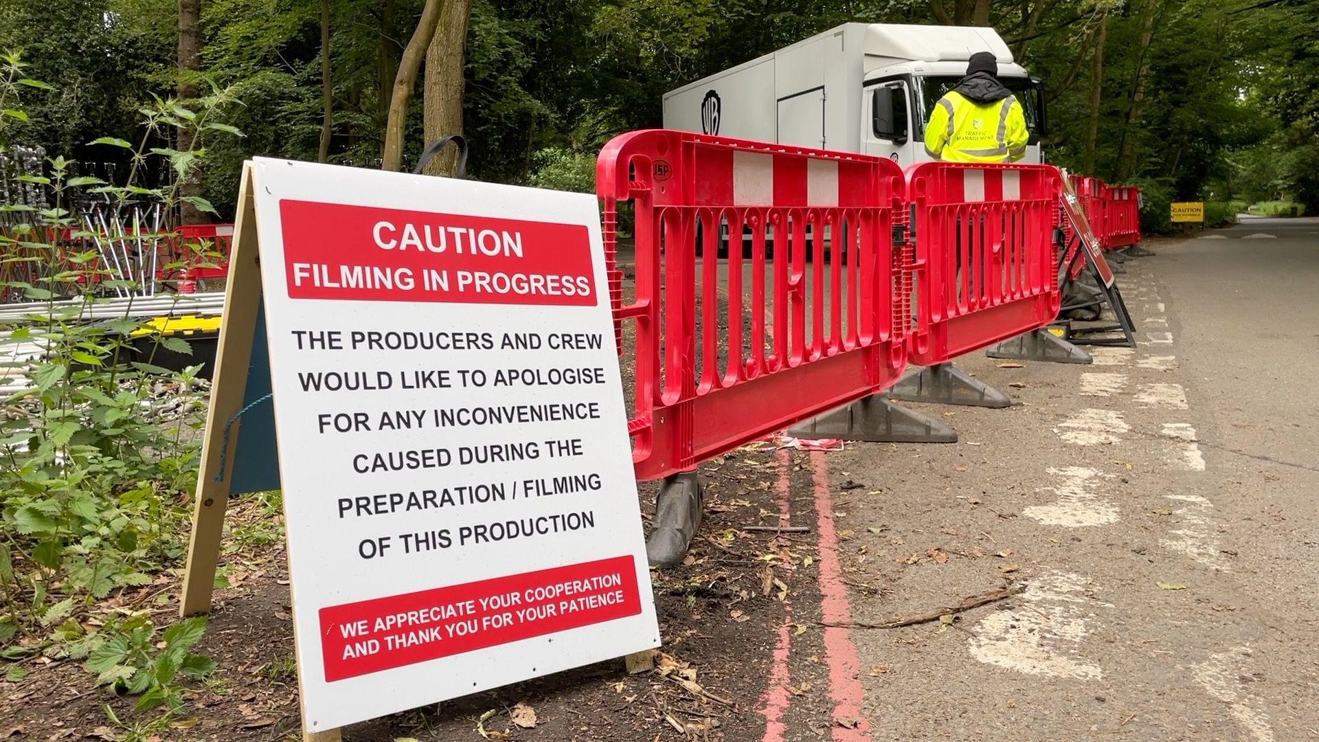 A white sign with a red banner and white writing is propped up on an A board, and stands next to a red plastic fence. A security guard wearing a high vis jacket stands behind the fence in front of a trailer. It is all at the edge of a road surrounded by trees. The sign reads: 'Caution. Filming in progress. The producers and crew would like to apologise for any inconvenience caused during the preparation/filming of this production. We appreciate your cooperation and thank you for your patience'.