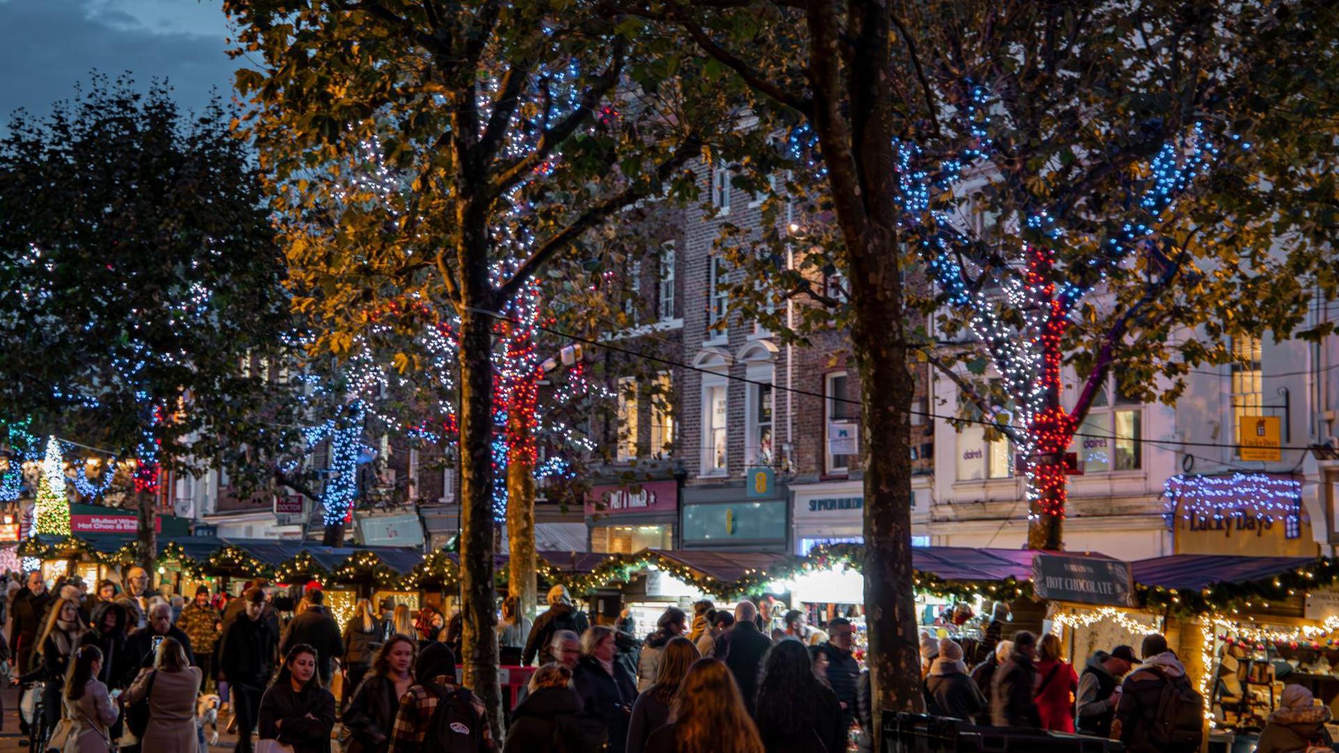 York's Parliament Street with Christmas decorations, a large crowd browsing craft and food stalls set up in small huts