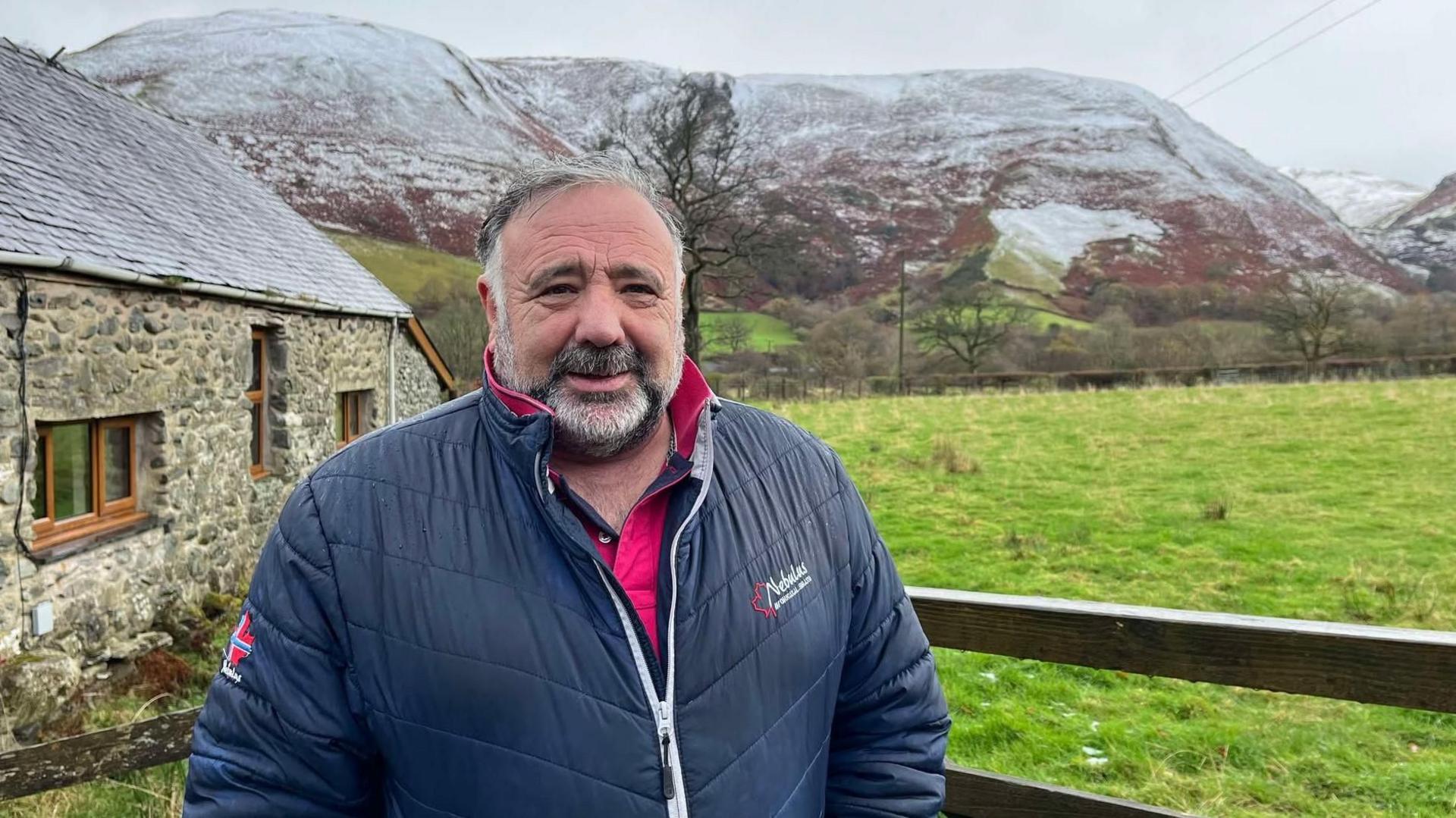 Councillor John Pughe Roberts, smiles at the camera, wearing a red polo shirt and a navy quilted jacket. The snow covered hills of Eryri national park stand behind him. 