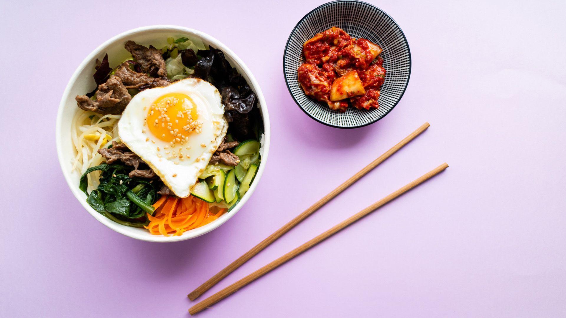 A bowl of beef bibimbap alongside a smaller bowl of kimchi and a set of wooden chopsticks on a light purple background 